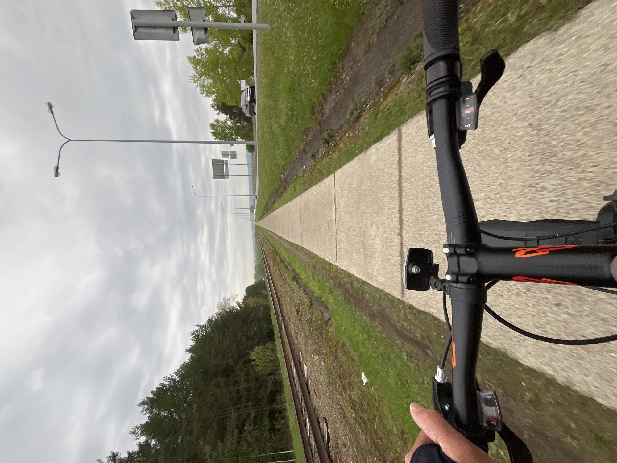 The image shows a first-person perspective from a bicycle rider on a paved path. To the left of the path are railroad tracks and a forest. To the right of the path is a grassy area alongside a road with streetlights. In the distance, there is water. The bicycle handlebars are visible in the foreground. The location is near Chaikovsky, Russia.