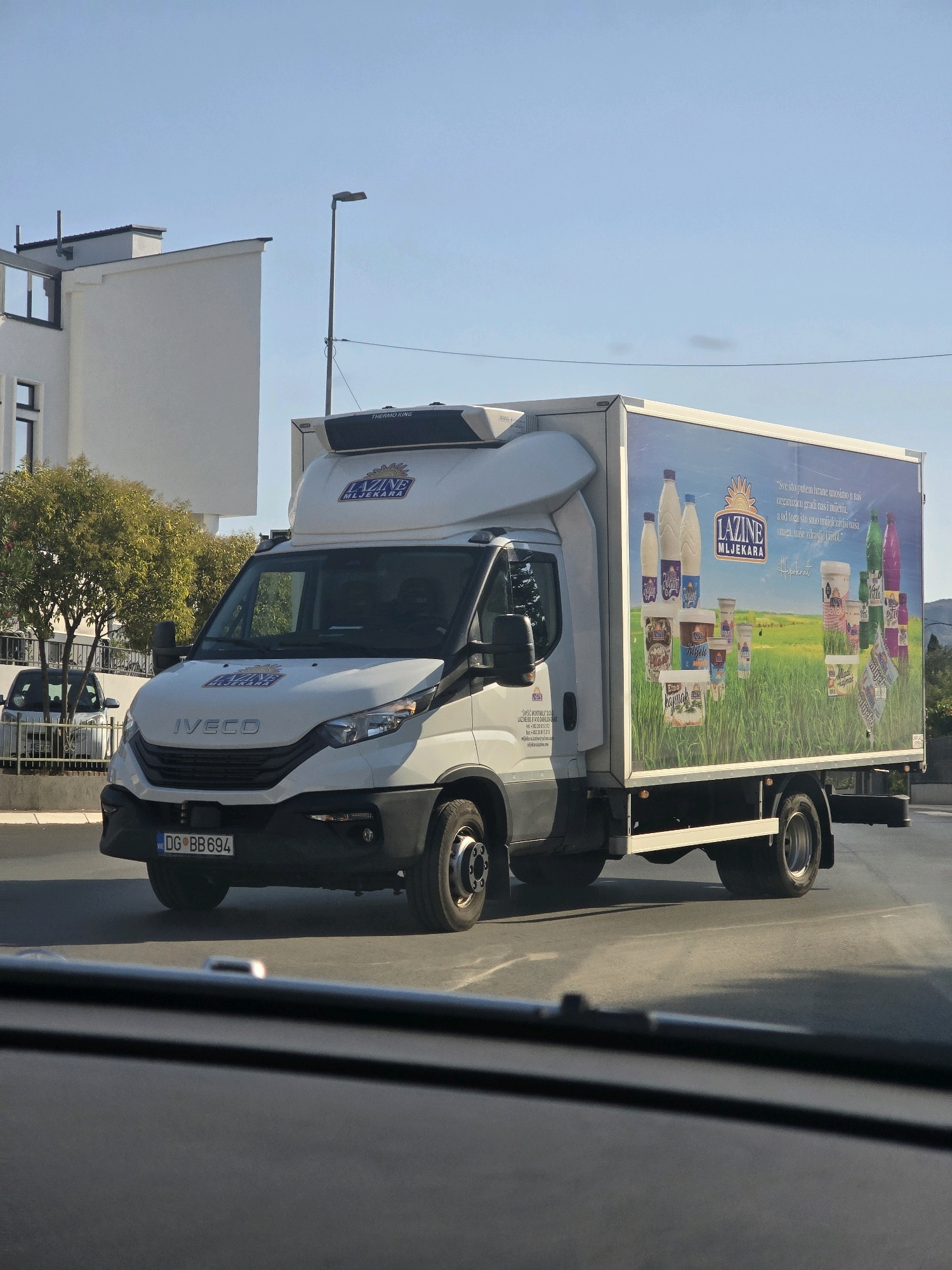 A white Iveco delivery truck with the "LAZINE MLJEKARA" logo is driving on a street in Podgorica, Montenegro. The truck has a large advertisement on its side depicting various dairy products against a green field and a blue sky. The license plate of the truck is DG BB 694. The truck is in motion, and its shadow is visible on the road. In the background, there is a modern white building and some trees. The sky is clear and blue. The photograph appears to be taken from inside another vehicle.