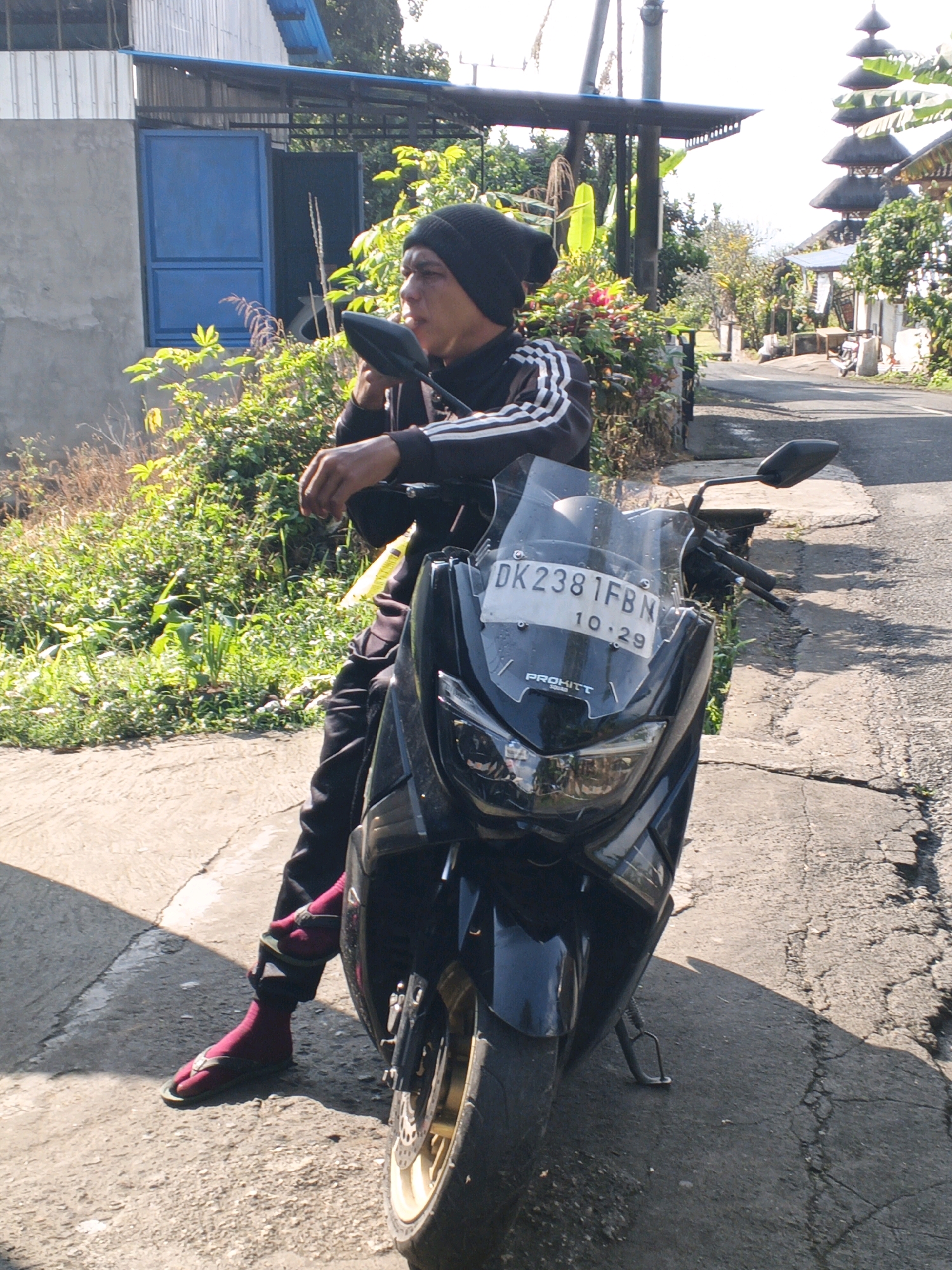 A man wearing a black beanie, black track jacket with white stripes, black pants, magenta socks, and dark sandals is sitting on a black motorcycle. The motorcycle has a clear windshield with a license plate displaying "DK 2381 FBN 10.29" and the text "PROHITT SQUAD". The man is holding onto the handlebars and looking to the right. The motorcycle is parked on a cracked asphalt road in front of lush green vegetation and a building with a grey concrete base and a corrugated metal upper section with a blue door. In the background, there are trees and structures, including what appears to be a tiered temple or pagoda-like building with a thatched roof. The scene is illuminated by sunlight, casting shadows on the ground. The location is identified as Penebel, Indonesia.