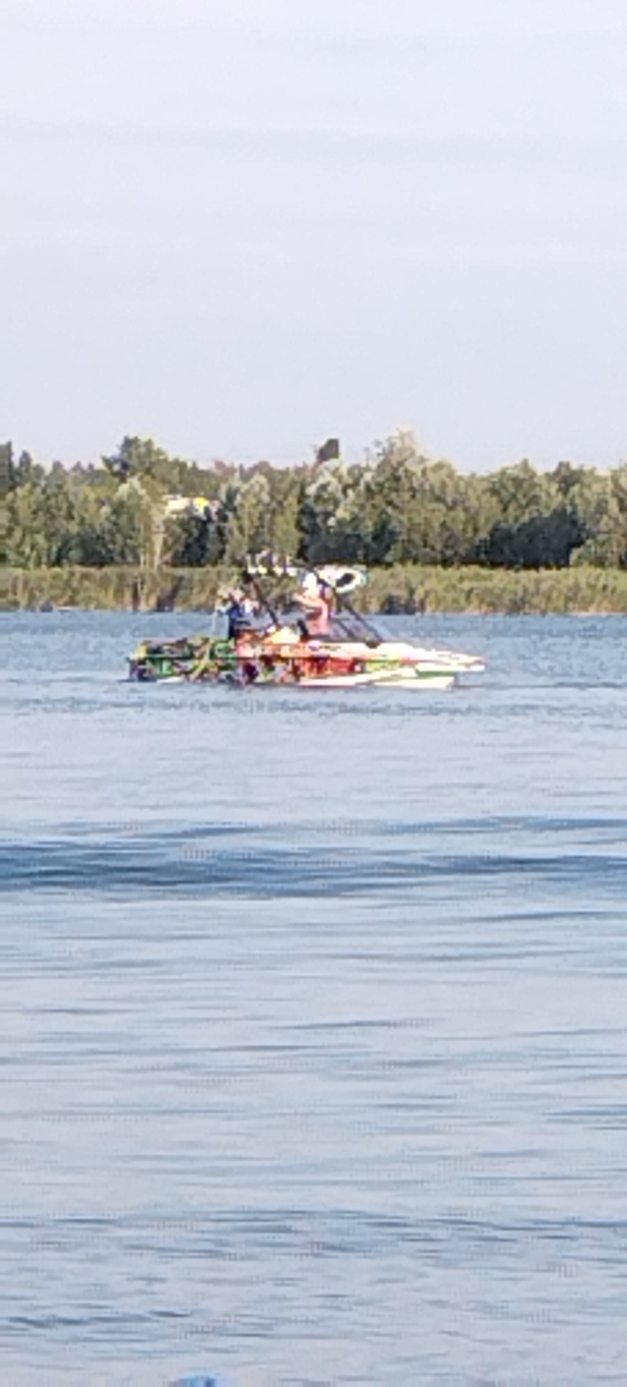 A brightly colored, modern speedboat with two individuals on board navigates a calm body of water, likely a river or lake. The boat features a vibrant, multi-colored pattern on its hull. One person, wearing a blue shirt, appears to be operating the vessel from the front, while another individual, in a pink shirt, sits behind. The surrounding environment consists of a tree-lined shore in the background, suggesting a natural or park-like setting within or near the city of Bezliudivka, Ukraine. The sky is overcast with light clouds.