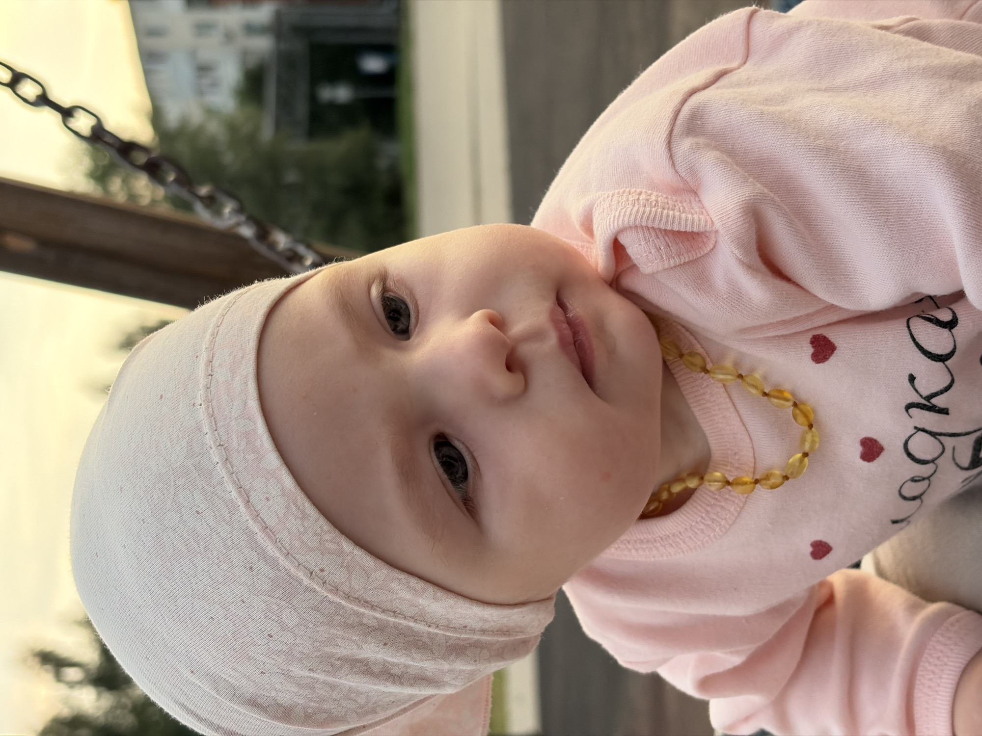 A close-up, eye-level, outdoor portrait captures a baby girl from the chest up, with her head turned to the right. She is wearing a light pink bonnet and a matching pink long-sleeved shirt adorned with small red heart graphics and text in black Cyrillic script. A beaded necklace, appearing to be amber, is around her neck. The background is softly blurred, showing the warm glow of sunset, some trees, and a chain swing set, suggesting the location is a playground. In the distance, a multi-story residential building is visible, typical of a Russian city. The overall impression is one of a peaceful, candid moment captured in a common urban setting.