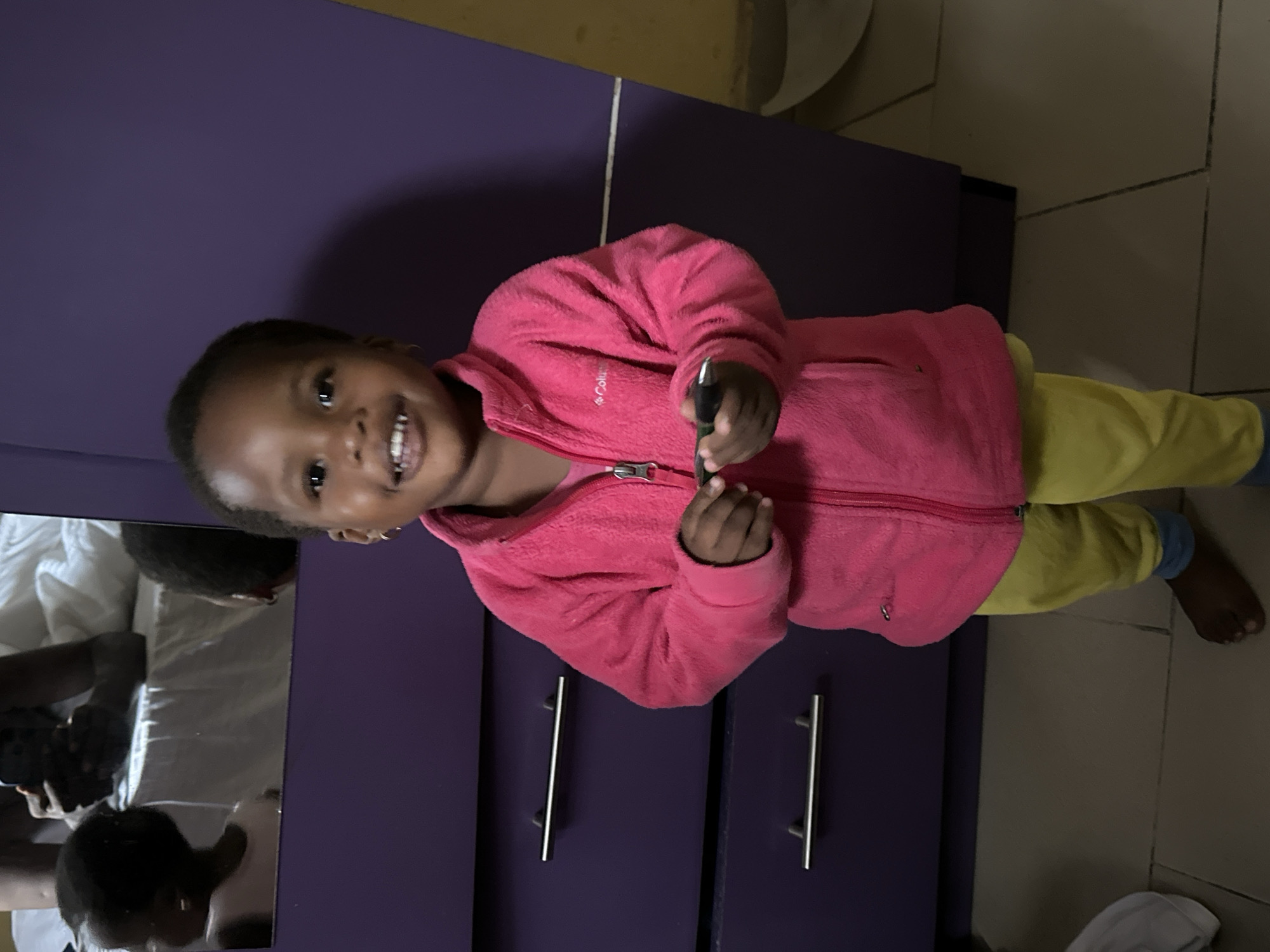 A young Black child, appearing to be between 2-4 years old, stands facing forward in a bright pink zip-up fleece jacket and yellow pants with light blue cuffs. The child has short, dark hair, brown eyes, and is smiling. In their right hand, they are holding a dark-colored object, possibly a pen or stylus. The child is positioned in front of a purple dresser with two silver drawer pulls. To the left of the child, a portion of a mirrored wardrobe is visible, reflecting the child's back and another person holding a smartphone. The floor appears to be tiled. The scene suggests an indoor setting, likely a bedroom or living area in Narayi, Nigeria, given the context.