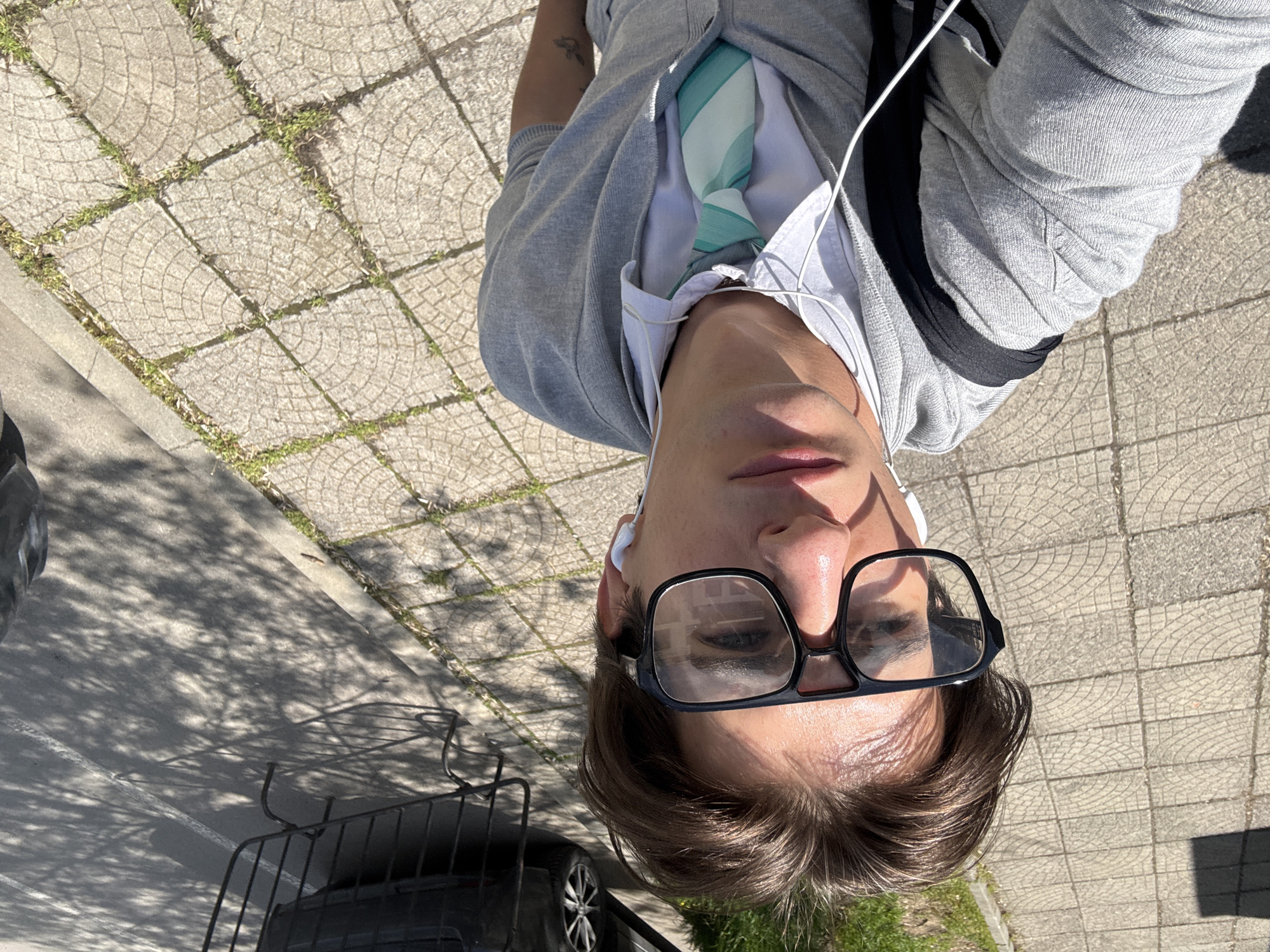 The image displays a person from an upward, inverted perspective, suggesting a selfie taken by holding the camera above their head. The subject is a young person with short brown hair, wearing black-framed glasses, a white shirt, a light blue and white striped tie, and a grey hooded jacket. White wired earbuds are visible in their ears. A small, faded tattoo, possibly a bird design, can be seen on their right arm. Their expression is neutral as they look directly into the camera.
The setting is an outdoor urban environment bathed in bright sunlight. The background features a paved sidewalk composed of square concrete tiles with a circular pattern, with grass growing in the cracks. Adjacent to the sidewalk is an asphalt road, partially obscured by dappled shadows cast by trees. A dark-colored car, including its wheel and tire, and a black metal structure, possibly part of a railing or car rack, are visible in the lower portion of the frame. The strong sunlight creates clear highlights and shadows, indicating midday or early afternoon. The scene is consistent with a street or park area in a city like Sofia, Bulgaria. No text is visible in the image.