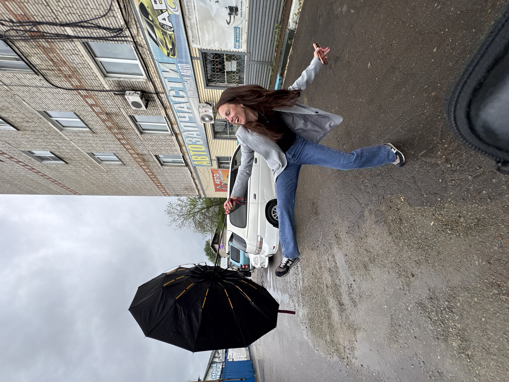 An outdoor photograph captures a joyous young woman in mid-action on a wet, paved street in Birobidzhan, Russia, under an overcast sky. The woman, with long dark brown hair, is dressed in a grey blazer over a black top, blue jeans, and sneakers. She wears a wide, beaming smile, with her arms outstretched; one hand holds a black umbrella with yellow ribs, which appears somewhat disheveled, possibly from wind or being in a state of opening/closing, and the other holds a smartphone. Her dynamic pose suggests she is jumping or playfully leaping.
In the background, a multi-story brick building runs along the left side of the frame, featuring multiple rectangular windows, visible electrical wires, and an air conditioning unit. A prominent commercial sign on the building advertises "АВТОЗАПЧАСТИ" (Auto Parts) and "ГРУЗОВЫХ АВТО" (Commercial Vehicles/Trucks), along with "АвтоСтиль." Further text on banners includes "RB-exide," "ЧИСТОЕ ДВИЖЕНИЕ!" (Clean Movement!), and "НАДЕЖНЫЕ" (Reliable). A white van is parked to the right of the woman, with a light blue vehicle visible further back. The ground is dark, wet asphalt, showing puddles, indicative of recent rain or damp weather. The overall scene depicts a moment of cheerful activity against an urban backdrop on a cloudy day.
