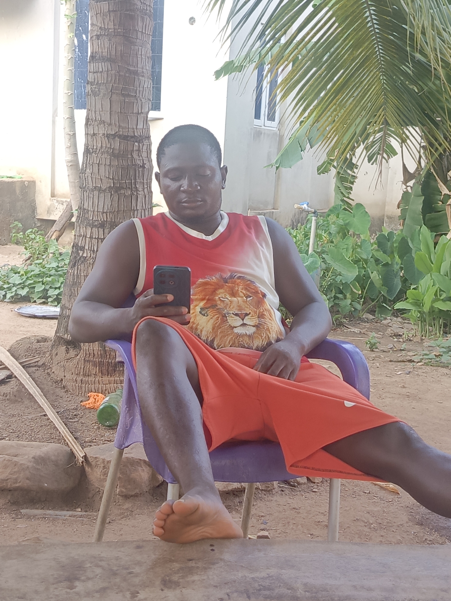An adult male sits in a purple plastic chair outdoors in Akwanga, Nigeria. The man has dark skin, a short haircut, and is wearing a tank top with a lion graphic and orange shorts. He is barefoot, looking at his smartphone, which he holds in his left hand. Behind him is the trunk of a palm tree and some vegetation. In the background, part of a building is visible. The ground appears to be sandy or dirt.