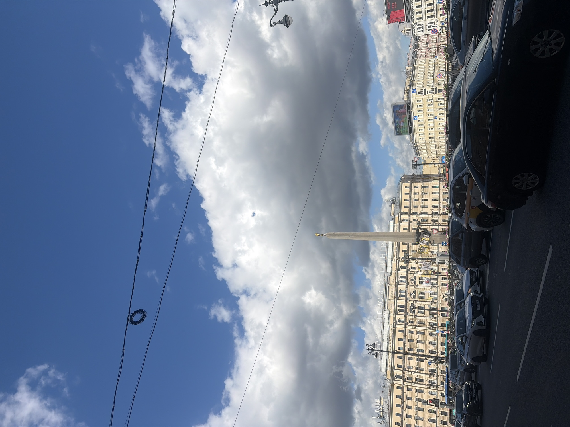The image captures a street scene in Saint Petersburg, Russia, likely near the Alexander Column, a prominent landmark. In the foreground, several cars are stopped at an intersection or in traffic on a paved road. The cars are a mix of sedans, SUVs, and a taxi. In the midground, a tall, slender obelisk, the Alexander Column, stands prominently, topped with a winged figure. Behind the column is a large, multi-story building with a beige facade, characterized by numerous windows and architectural details. The building appears to be an older, historic structure. Further in the background, more buildings of similar architectural style line the street, interspersed with some advertisements. A streetlamp with an ornate design is visible to the left of the column. The sky above is a mix of bright blue and large, dramatic white and grey clouds, suggesting a partly cloudy day. Overhead, electrical wires are strung across the frame, with a coiled bundle of wires hanging from one of them. The overall impression is that of a busy urban environment on a day with dynamic weather conditions.