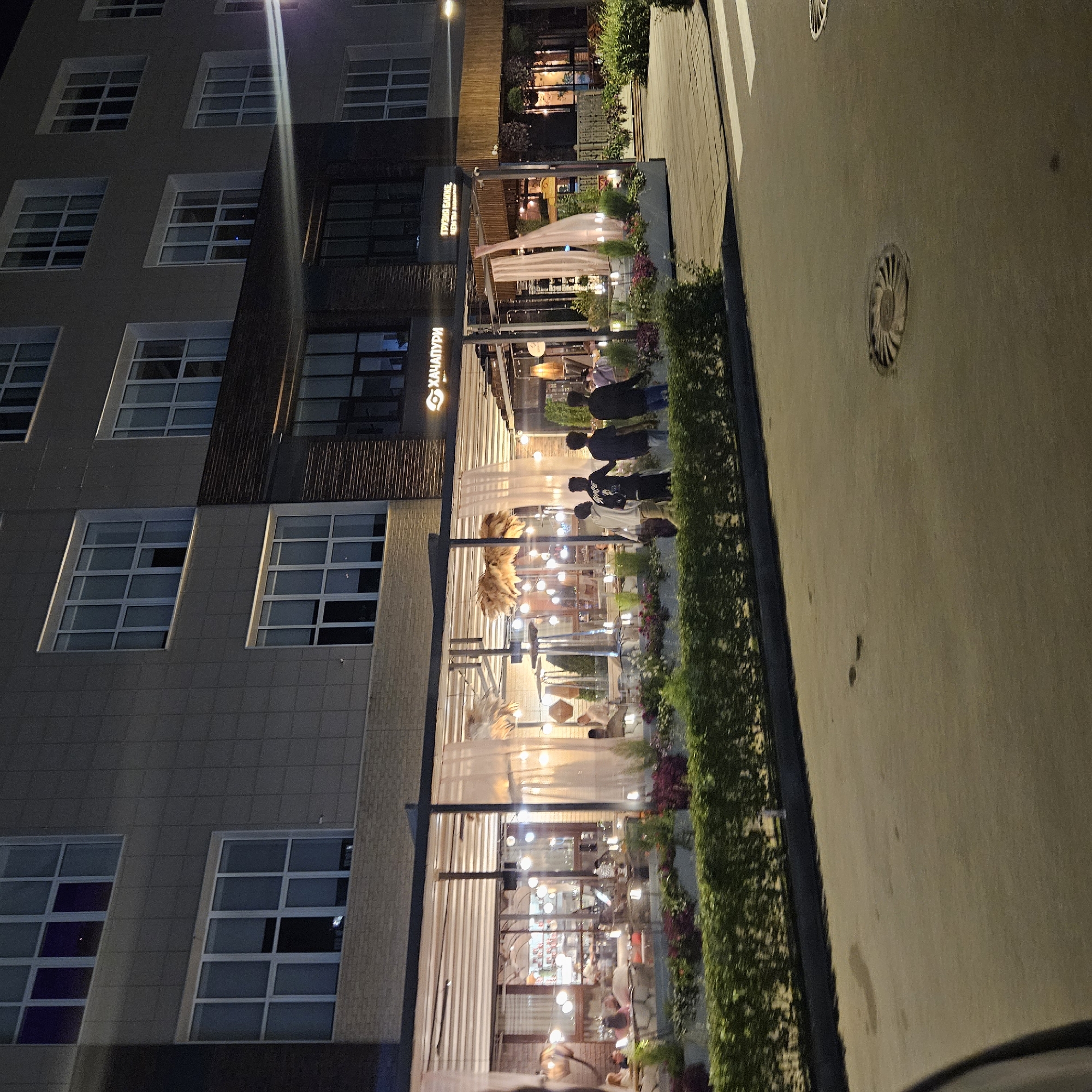 A nighttime street-level exterior shot shows the facade of a modern building with multiple floors of windows. The ground floor features a brightly lit outdoor seating area of a restaurant, with white sheer curtains and hanging decorative lights. Several people are seated at tables within the restaurant, and a few individuals are walking outside on the sidewalk. The building has a sign that reads "ХАЧАПУРИ" in Cyrillic script, identifying it as a Georgian restaurant. The street in front of the establishment is paved, with a crosswalk visible. A dense green hedge runs along the sidewalk.