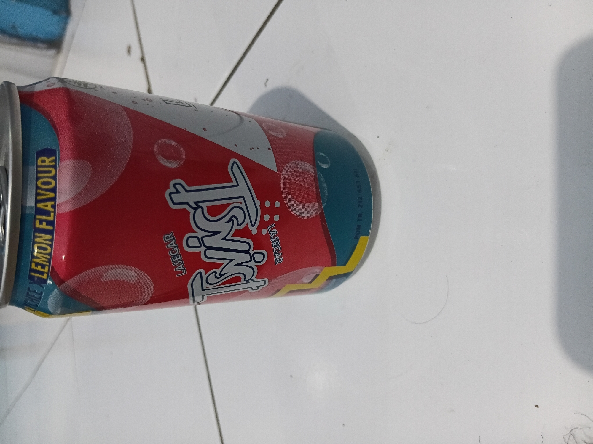 A close-up, eye-level shot shows a red and blue soda can with the brand name "TWIST" in large, stylized white lettering. The can is positioned on a white tiled surface. The top portion of the can has a label that reads "LYCHEE X LEMON FLAVOUR." Below the brand name, the word "LASEGAR" is printed vertically in small white text. The bottom of the can features a teal band with a registration number "POM TR. 212 653 611" printed in black. The lighting is somewhat dim, casting a shadow from the can onto the tiled floor. The environment appears to be indoors, possibly a kitchen or a shop, based on the tiled surface, which is common in Indonesian households and businesses. The specific location within Tanggul Wetan, Indonesia cannot be determined from the image alone, as it lacks any identifiable landmarks or context clues specific to that area.