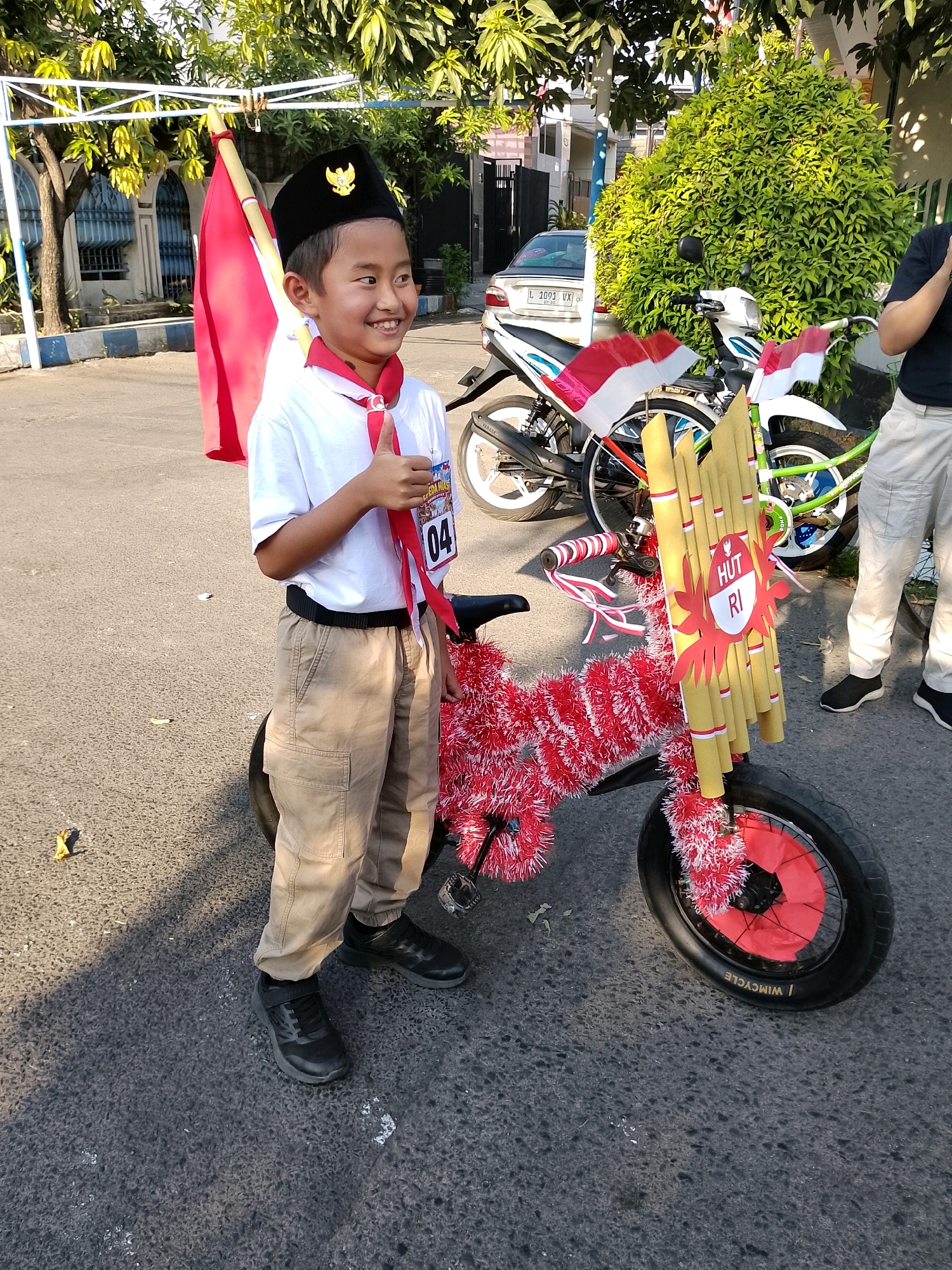 A young Indonesian boy, dressed in a white shirt, tan pants, a red sash, and a black peci hat with a gold emblem, stands next to a decorated bicycle. The bicycle is adorned with red tinsel, Indonesian flags, and a decoration made of bamboo tubes with a red and white sign that reads "HUT RI." The boy is giving a thumbs-up gesture. In the background, a white motorcycle with Indonesian flags is parked, along with a light-colored car and a lush green bush. The scene appears to be on a paved street, possibly during a parade or event, with trees and buildings visible in the distance. The overall impression is a festive and patriotic atmosphere.