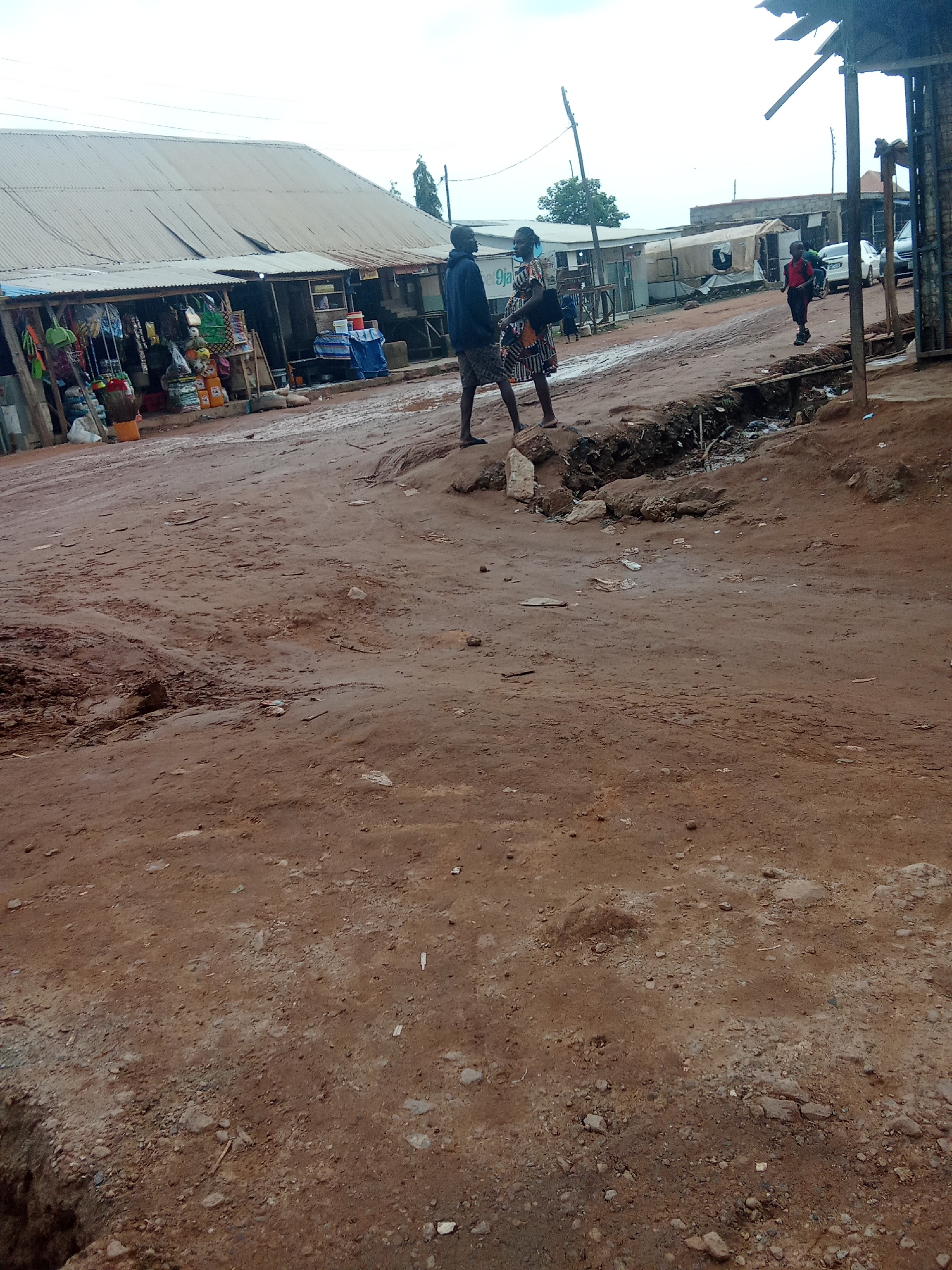 In a medium-wide shot, a dusty, unpaved road occupies the foreground of the image, with a large puddle or muddy area to the left. Shops with corrugated metal roofs line the right side of the road. A man in a dark shirt and shorts stands on the right side of the road, facing left. Further down the road, a woman in a patterned dress walks away from the camera, and another person, possibly a child, wearing a red shirt and dark shorts, walks towards the camera. In the background, several vehicles are parked, and more shops or stalls are visible. The sky is overcast.
