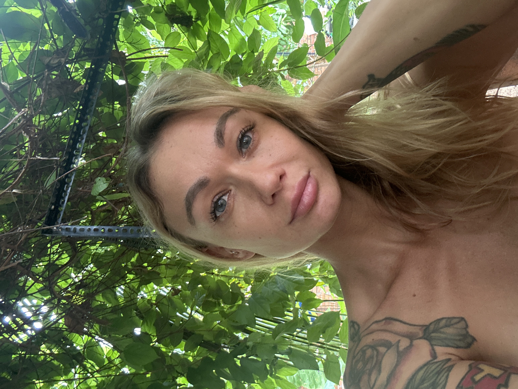 A fair-skinned woman with light hair and visible tattoos is the primary subject of the image, taken from a low angle, seemingly looking up at her. She appears to be outdoors, surrounded by lush green foliage and what looks like a metal trellis or support structure. The location is likely Mũi Né, Vietnam, based on the tropical vegetation. There is no further evidence in the image to support any other details related to Mũi Né or the woman's activities.