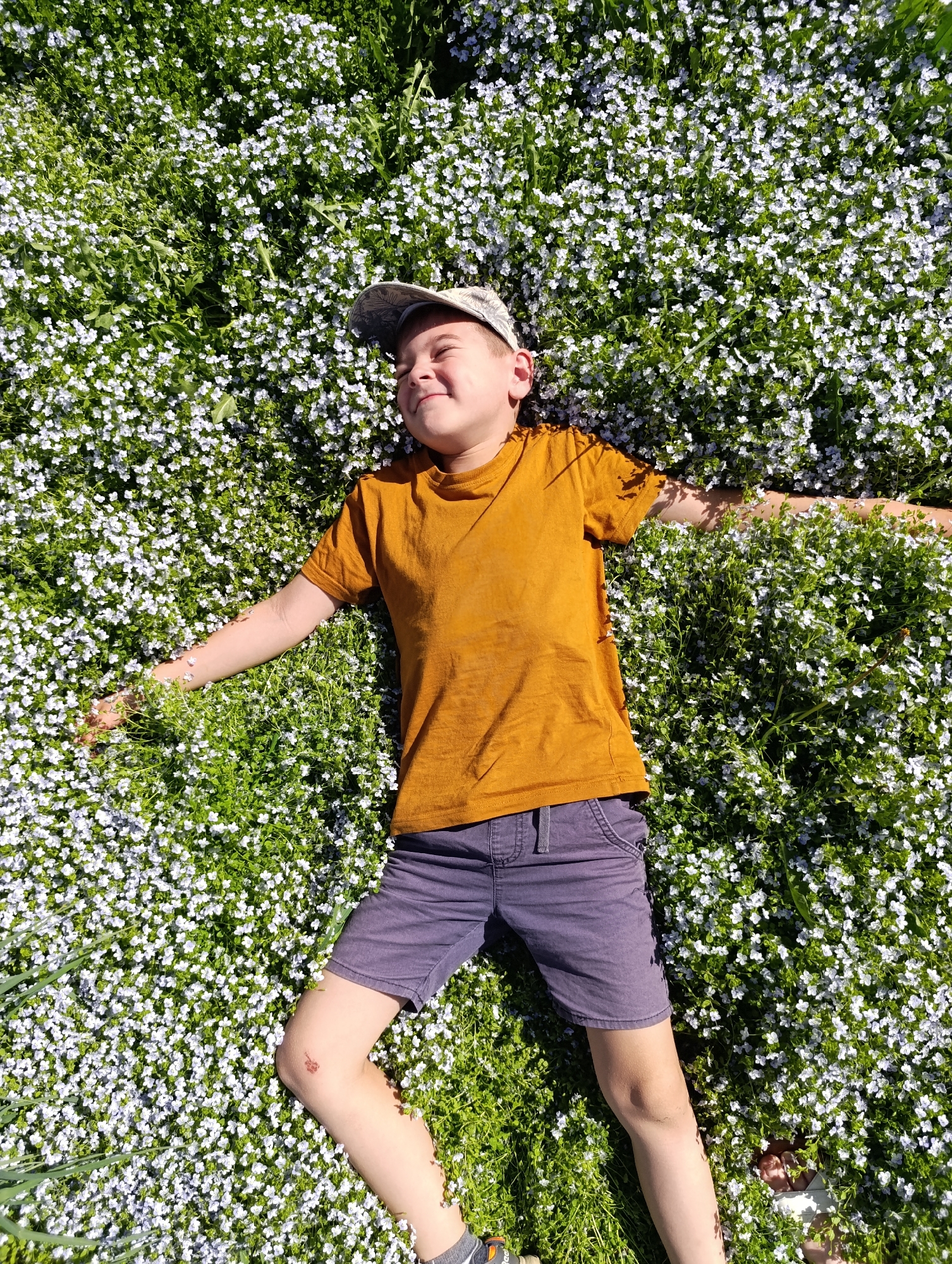 A boy lies in a field of small white flowers. He is wearing a mustard yellow t-shirt, gray shorts, a patterned baseball cap, and sneakers. His arms are outstretched. He has a small abrasion on his left knee. The location is Chaikovsky, Russia.