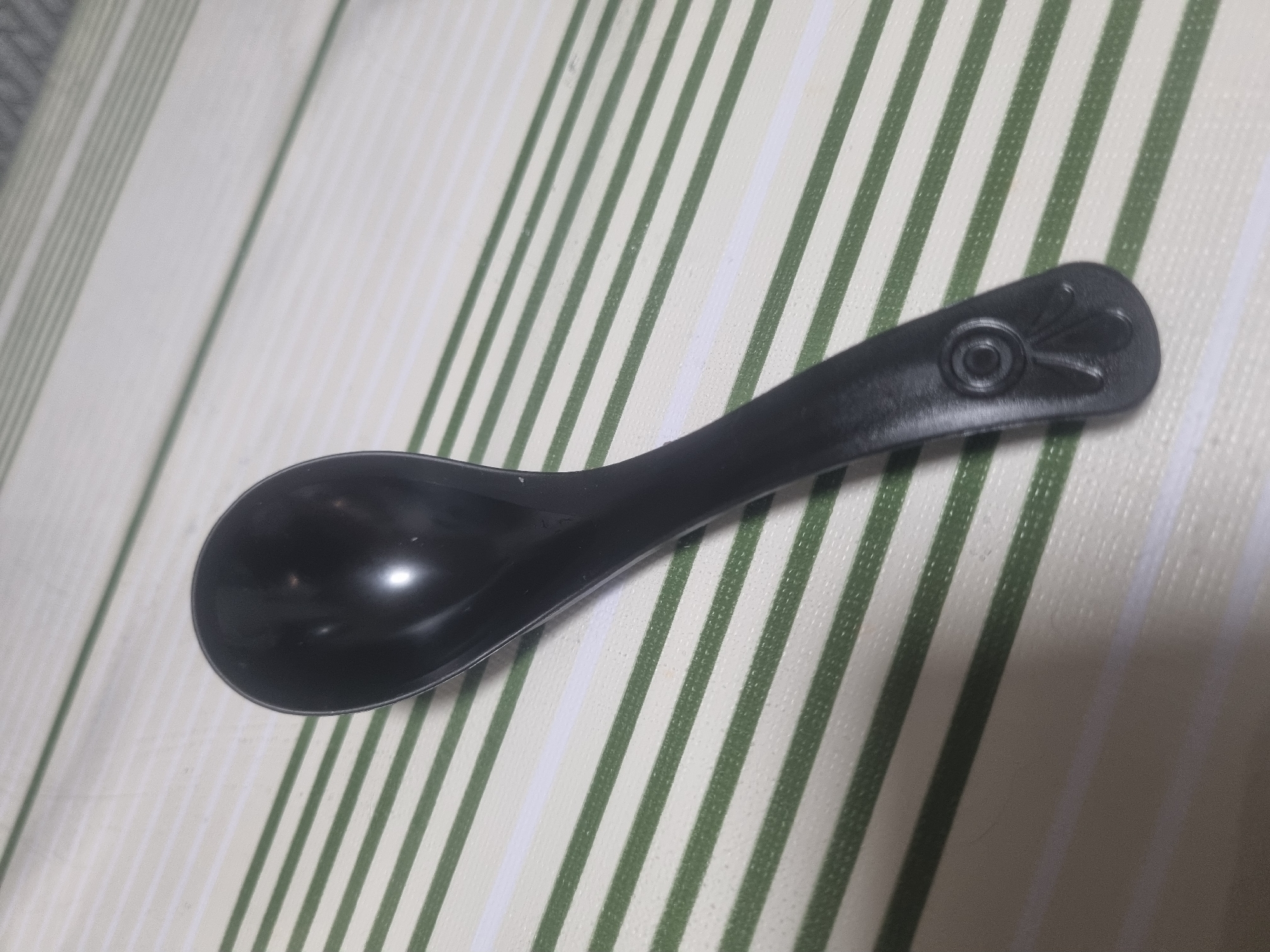 A black plastic spoon rests diagonally on a striped surface. The spoon's bowl is at the upper left, facing upwards, and its handle extends towards the lower right. The handle has a decorative element at its end, featuring a circular indentation with a petal-like design below it. The background consists of alternating horizontal stripes of light green and white. The image does not contain any information about the city of Troitskoye, Russia.