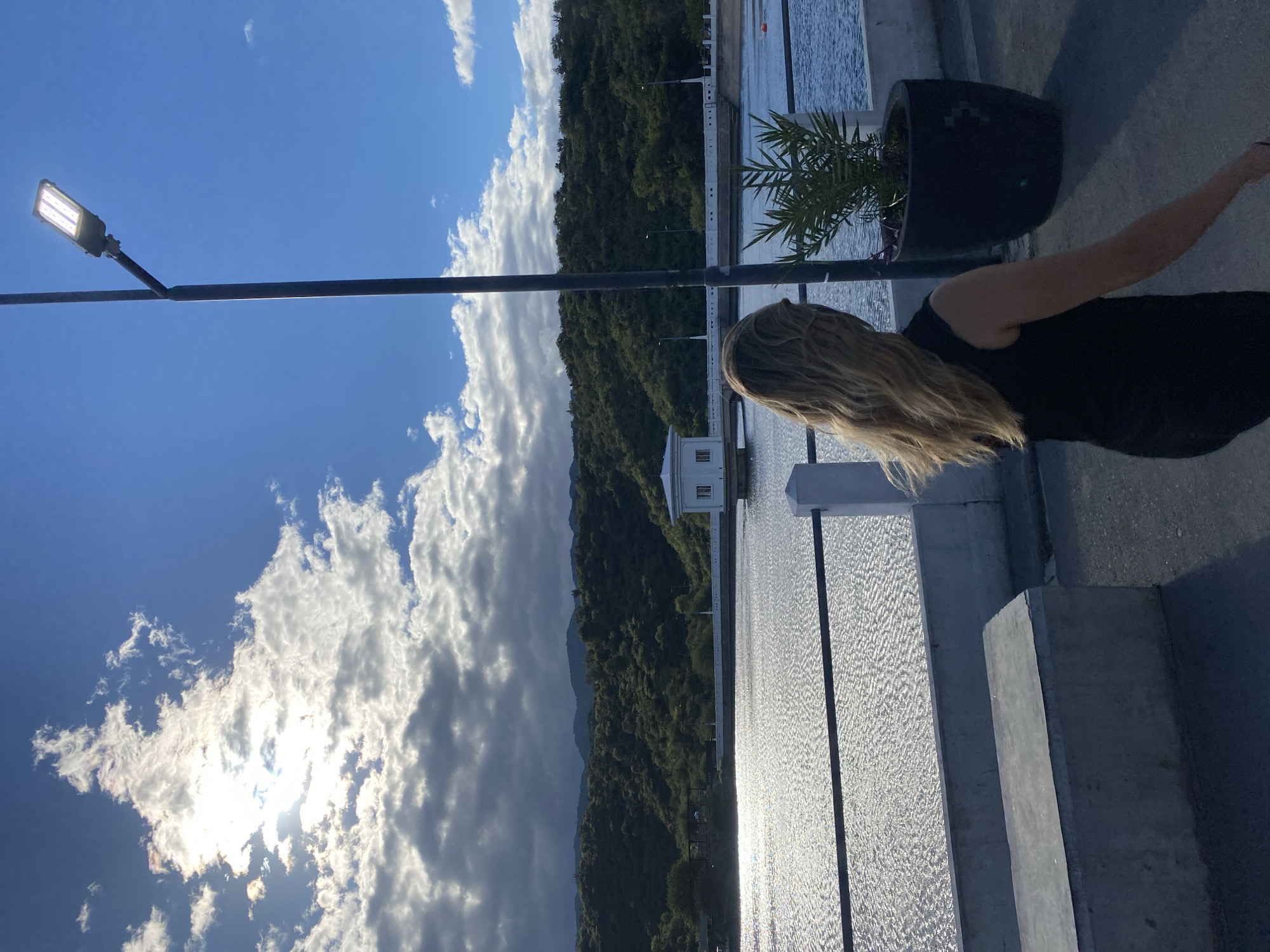 A woman stands on a concrete structure alongside a body of water, possibly a reservoir or lake, near San Fernando del Valle de Catamarca, Argentina. She is wearing a black sleeveless top. A black potted plant sits behind her. A small white building is seen across the water. A densely forested hillside is across the water. The sky is bright with scattered clouds. A tall black lamp post stands to the left. The sun is shining through the clouds.