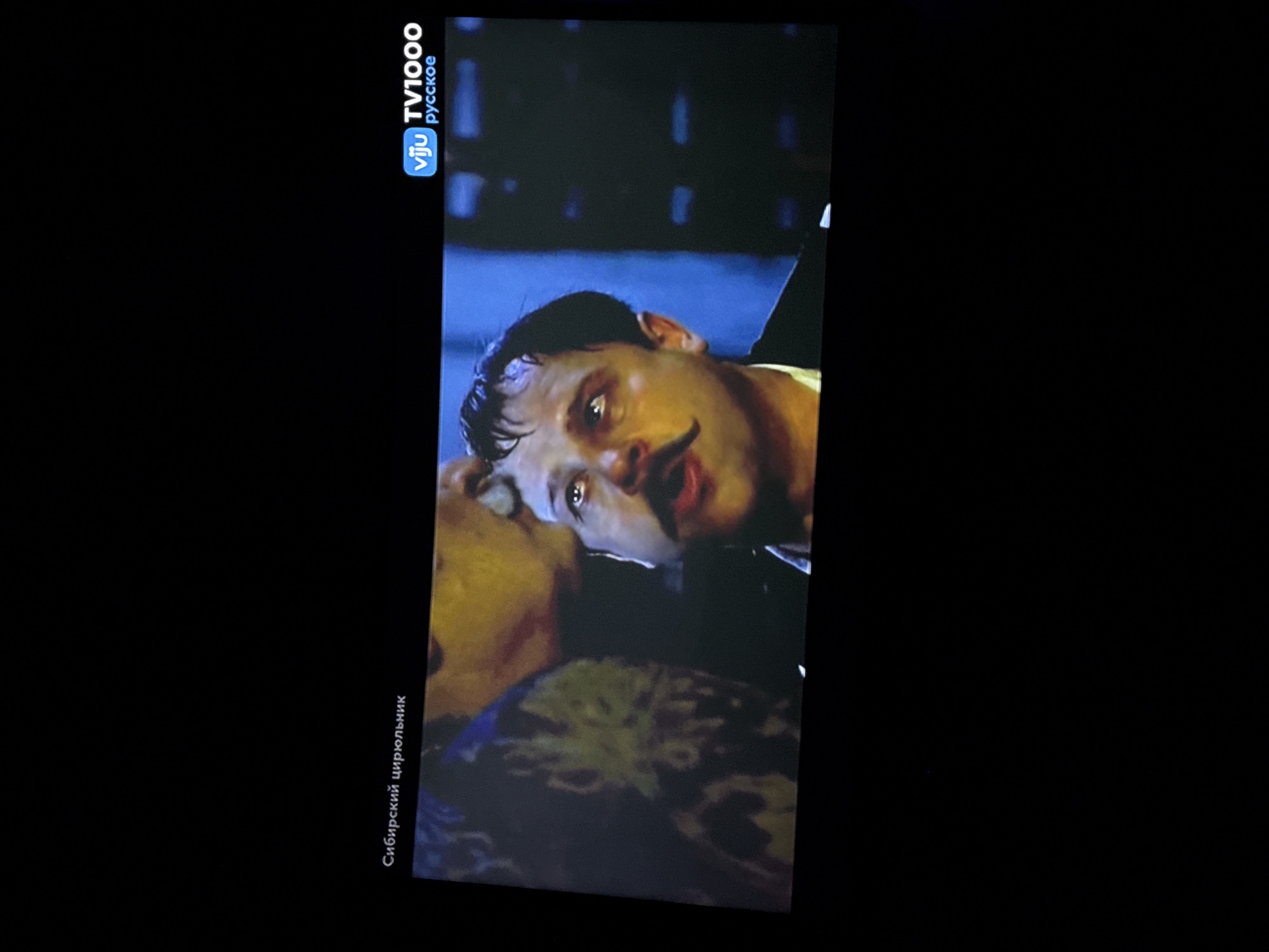 The image shows a close-up of a man's face with a prominent mustache, looking upwards with his mouth open. He appears to be in distress or surprise. The background is dark and blurry, with some indistinct shapes and colors. In the top left corner, there is text in Russian that reads "Сибирский цирюльник" which translates to "The Siberian Barber". In the top right corner, there is a logo for "vju TV1000 русское". The scene is likely from a movie or television program. There is no visible indication of the city of Kokkorevo, Russia in the image.