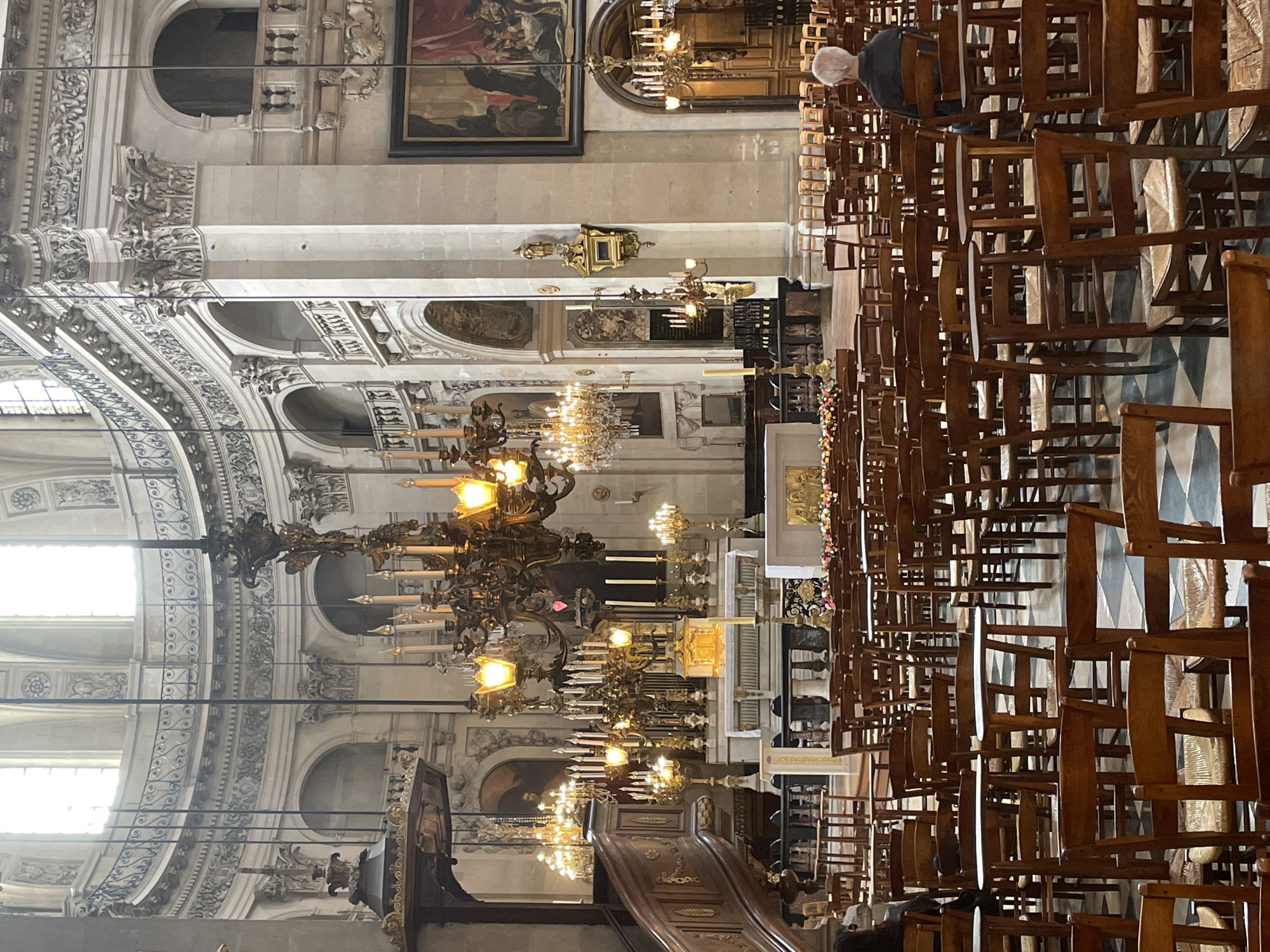 The image shows the interior of the Saint-Sulpice Church in Paris, France. The vast, opulent space features high ceilings with ornate architectural details, including columns, arches, and a large dome. Several large, antique chandeliers with many candles hang from the ceiling.
In the foreground, rows of wooden chairs with woven seats are arranged on a checkered floor. Some chairs are empty, while a few people are seated. A man with gray hair, facing away from the camera, is seated in one of the chairs in the middle ground. A woman with dark hair is visible on the far left, also seated.
Further into the church, a large wooden pulpit is situated to the left. The altar area is visible in the background, adorned with flowers and religious artifacts. A large painting hangs on the wall to the right. The overall ambiance is one of grandeur and solemnity, characteristic of a historic Parisian church.