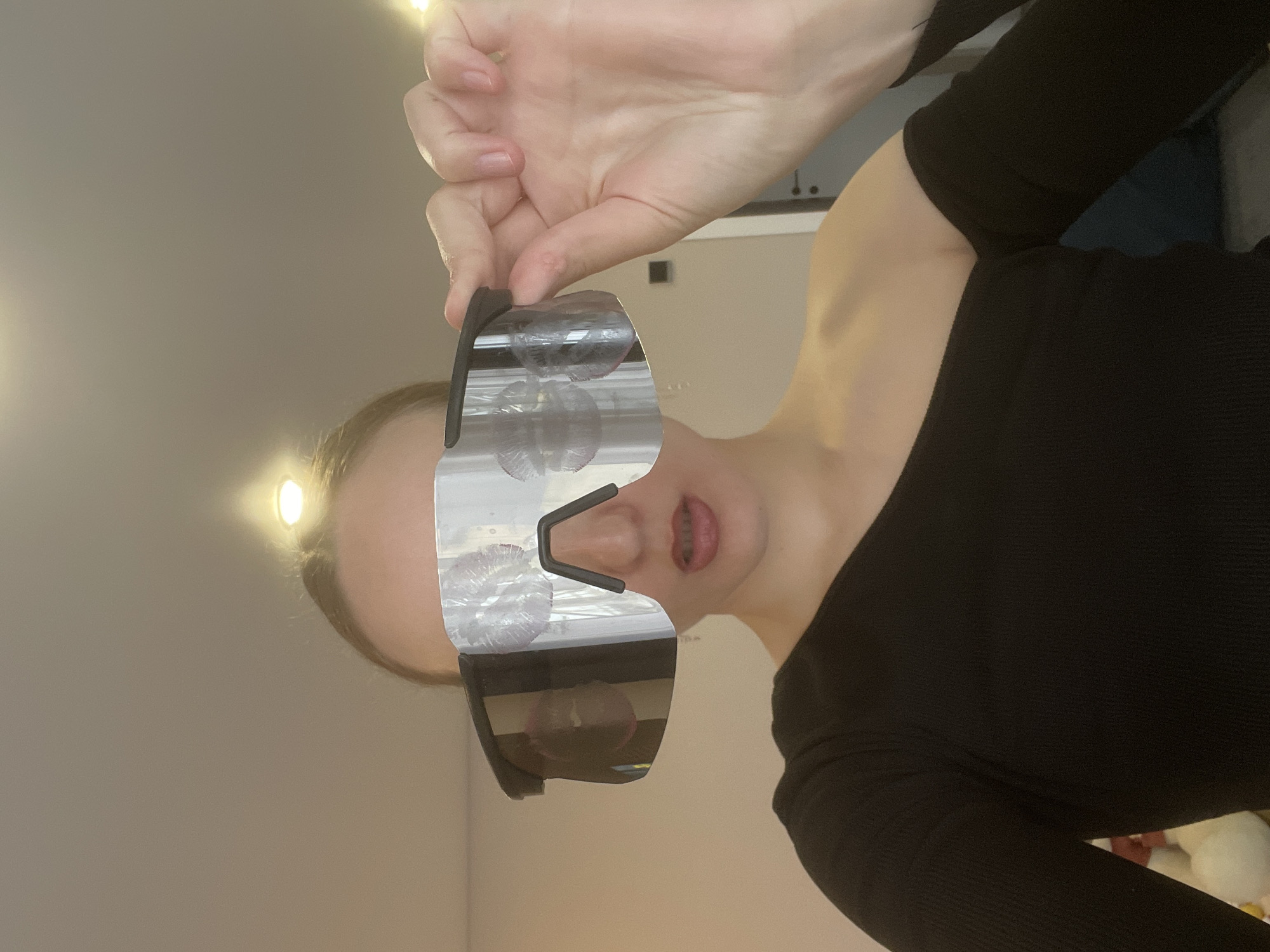 A young person is depicted indoors, holding a pair of large, mirrored sunglasses up to their face. The subject, likely female, wears a black, long-sleeved top with a ribbed texture. Their hands are visible, gripping the top edge of the oversized, futuristic-style eyewear.
The most notable detail of the sunglasses is the presence of several light-colored lipstick kiss marks scattered across the reflective lenses, indicating a playful interaction. Below the glasses, the person's lower face is visible, showing slightly parted lips with what appears to be dental braces on the lower teeth. Their expression is neutral.
The background is a plain, light-colored wall or ceiling, suggesting a simple indoor environment. Two bright, warm-toned light sources are visible in the background, creating a soft, warm glow. The lighting suggests an artificial indoor setting, making the time of day ambiguous. No text is visible in the image. The scene takes place in Podshivalovo, Russia.