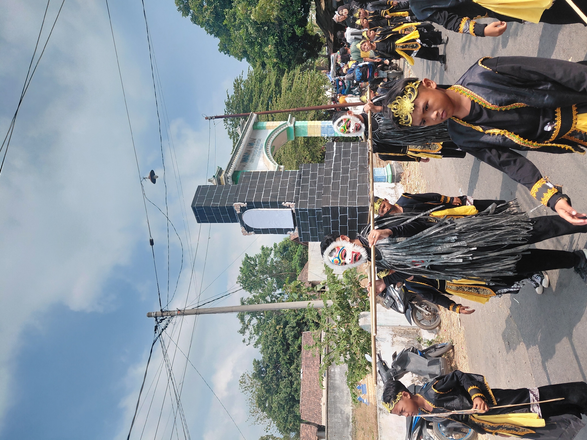 This image captures a procession taking place on a paved street in Bimo, Indonesia, under a partly cloudy sky. In the foreground, several young boys in traditional black and yellow costumes are marching. Some wear ornate gold headbands, and one boy in the front left carries a wooden staff. Behind him, other boys are dressed in black outfits adorned with yellow accents and fringed black decorations around their necks. Several of them wear colorful masks with white bases, red and green eye details, and white fringe around their heads. They are carrying a long bamboo pole horizontally, indicating they may be carrying something unseen or participating in a symbolic carrying ritual.
Further down the street, more participants in similar attire are visible, as well as other people watching the procession. Behind the marchers, a large, dark, brick-like monument stands prominently, appearing to be made of dark grey or black bricks with some lighter detailing. An archway structure with the inscription "MAKAM UMUM" is visible beyond the monument, suggesting the procession is passing through or near a public cemetery or significant historical site. Motorcycles are parked on the side of the road, and residential buildings with tiled roofs are visible in the background. Green trees line the area, and utility poles with numerous wires are present. The overall scene suggests a cultural or religious event taking place in a semi-urban or village setting in Bimo, Indonesia.