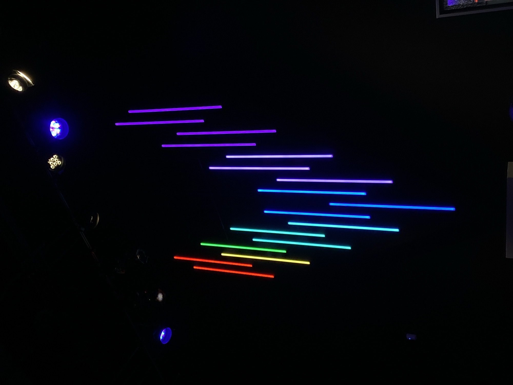 The image shows a dark room with a series of brightly colored vertical neon lights hanging from the ceiling. The lights are arranged in a V-shape, with red, orange, yellow, green, cyan, blue, and purple lights forming the sequence from left to right. Above the lights, several spotlights are visible, including a blue spotlight on the left, a cluster of yellow lights in the center, and a white spotlight on the upper right. The background is entirely black, creating a dramatic contrast with the illuminated lights. The scene appears to be an indoor installation, possibly in a museum, art gallery, or entertainment venue. There are no people or specific landmarks visible that would indicate a precise location within Washington, United States of America.