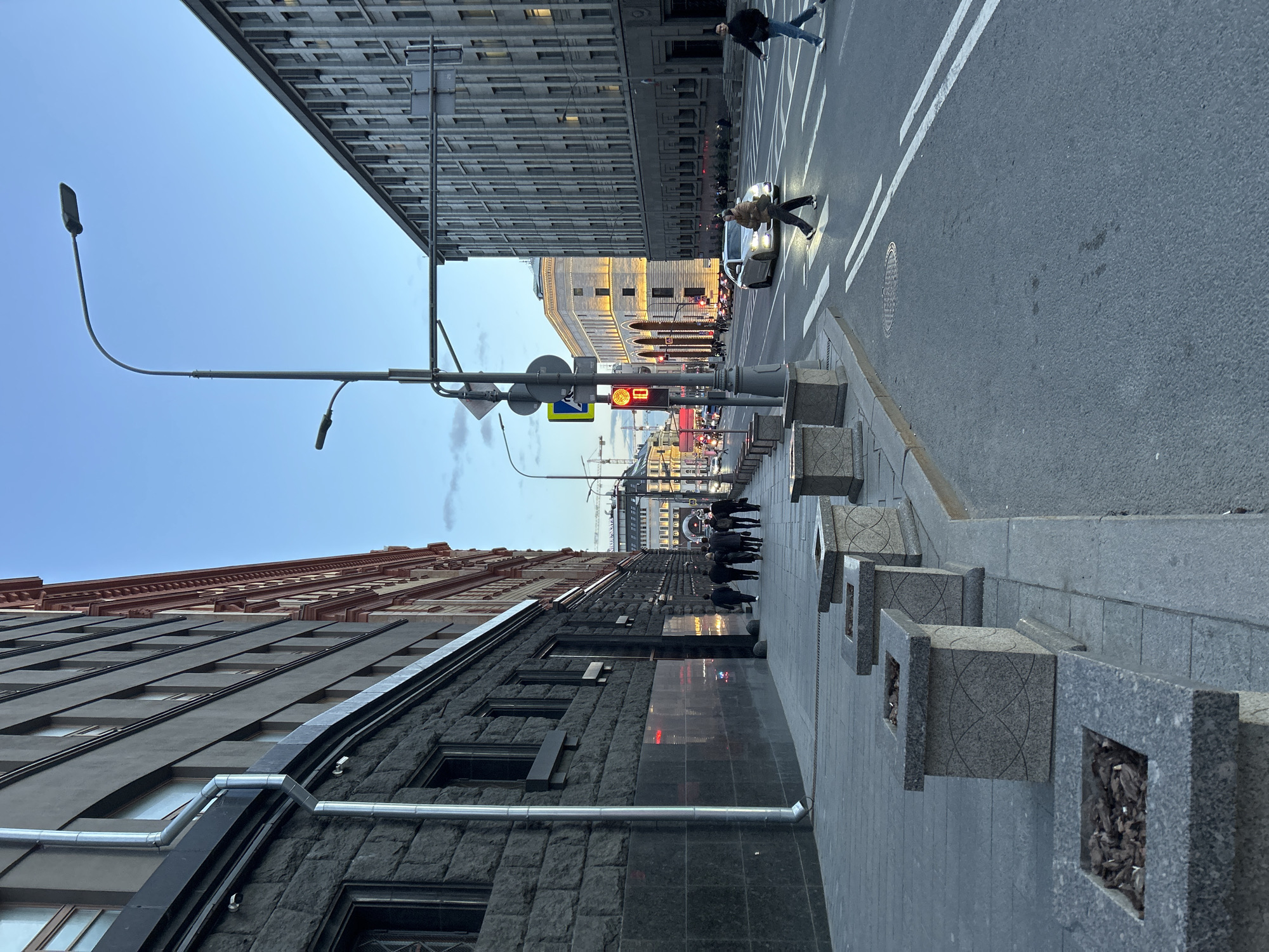 This is an urban street scene captured during dusk or early evening, as indicated by the soft, ambient light and the activated artificial lights. The image depicts a city street extending into the background, flanked by multi-story buildings on both sides.
In the foreground on the left, a tall building with dark, rough-textured stone cladding dominates the lower half, featuring multiple windows and a metal drainage pipe running vertically. Above this, the upper stories of an older, lighter-colored building with ornate architectural details are visible. On the right, a wide paved sidewalk features a series of square, grey stone planters or barriers with decorative patterns, some containing dry leaves or debris.
The street, which appears to be asphalt, includes a white-striped crosswalk. A silver or light-colored car is stopped on this crosswalk, with its headlights on. A person is seen walking across the street in front of the car, and another individual is further along the crosswalk. Further down the street, a group of people are gathered on the sidewalk.
Overhead, several streetlights are visible, including one with a distinctive curved arm. A red traffic light, clearly displaying the number "10" (likely a countdown), hangs above the street. Nearby, a street sign with blue and yellow colors shows a white arrow and a pedestrian icon. In the distance, lights from other buildings and street infrastructure illuminate the streetscape. The sky above is a clear, light blue, gradually fading towards the horizon. The architectural styles of the buildings suggest a mix of contemporary and historical structures.