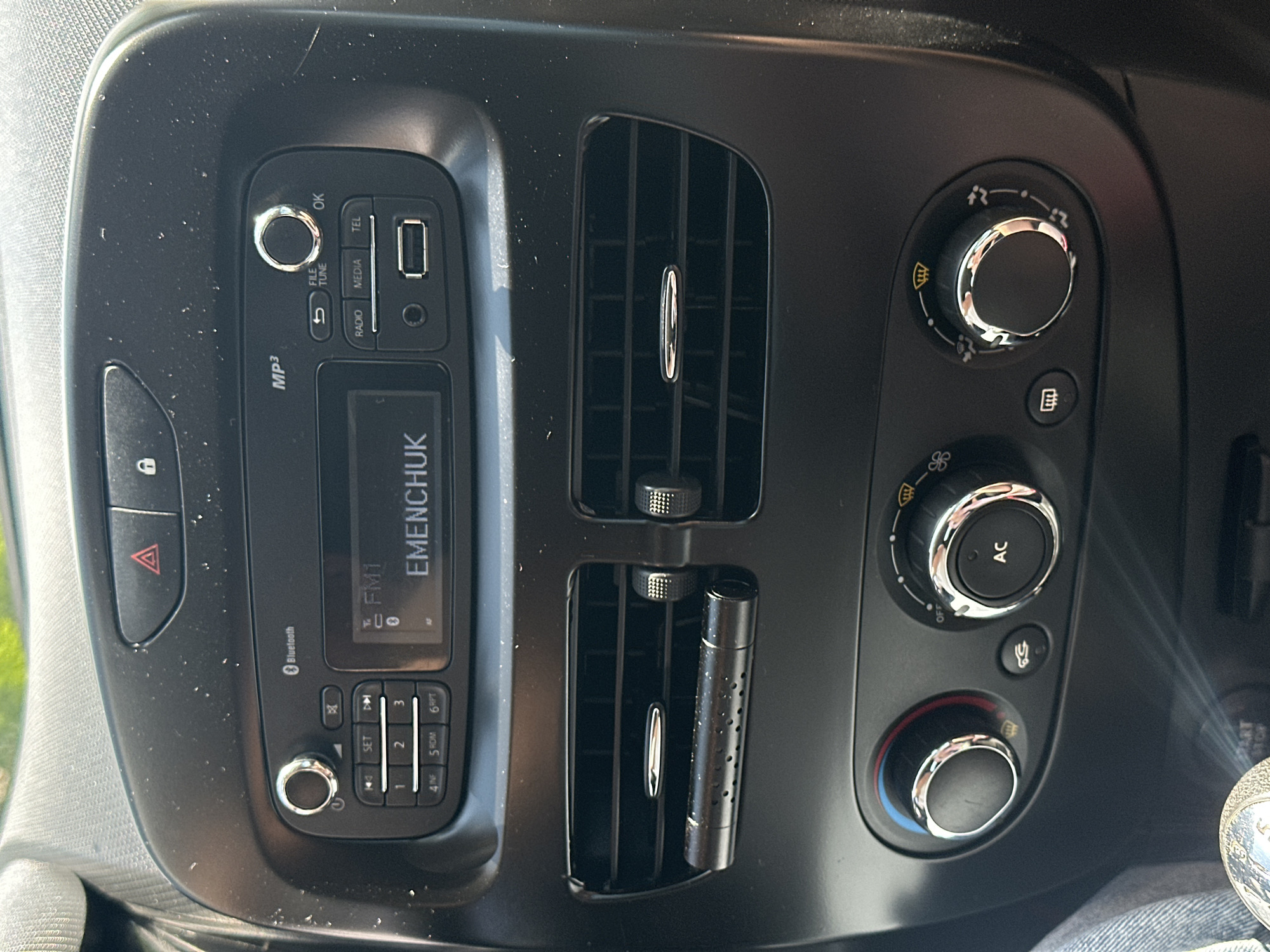 The image shows the interior of a car, specifically the dashboard area. A car's audio system with a display screen is prominently featured, showing "FM1" and "EMENCHUK". There are various buttons and dials for controlling the radio, media, and phone functions. Below the audio system are two air vents, and further down are climate control dials, one of which is labeled "AC". The dashboard appears dusty. The surrounding environment outside the car is not visible. The image was taken in Mala Kokhnivka, Ukraine.