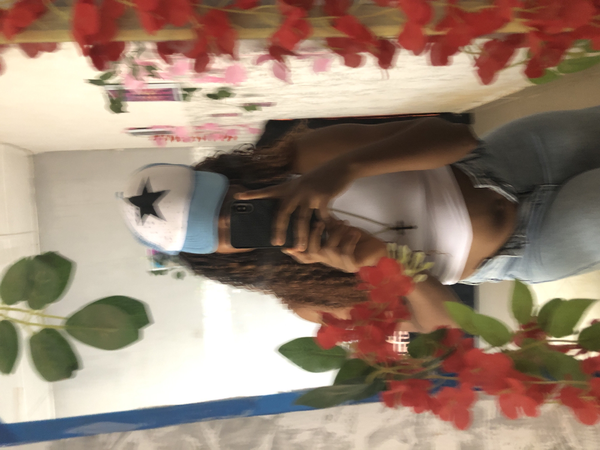 A person is taking a selfie in a mirror. The person is wearing a white tank top and light blue jeans, and a hat with a black star on it. The mirror is decorated with red flowers and green leaves. There is a plant with green leaves on the left side of the mirror. The location is not discernible from the image, so I cannot confirm if it is in Kashere, Nigeria.