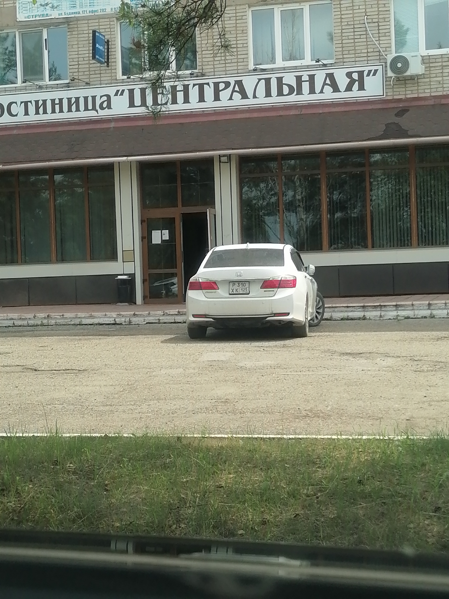 The image captures the facade of a brick building, likely a hotel given the signage that reads "Гостиница 'Центральная'" (Hotel 'Centralnaya'). The building features large windows with dark frames and an entrance with glass doors. A white sedan is parked in front of the building, partially obscuring the lower portion of the entrance. An air conditioning unit is visible on the upper right side of the building. The area in front of the building appears to be a dirt or gravel lot, with a strip of green grass bordering it. The location is identified as Lesozavodsk, Russia.