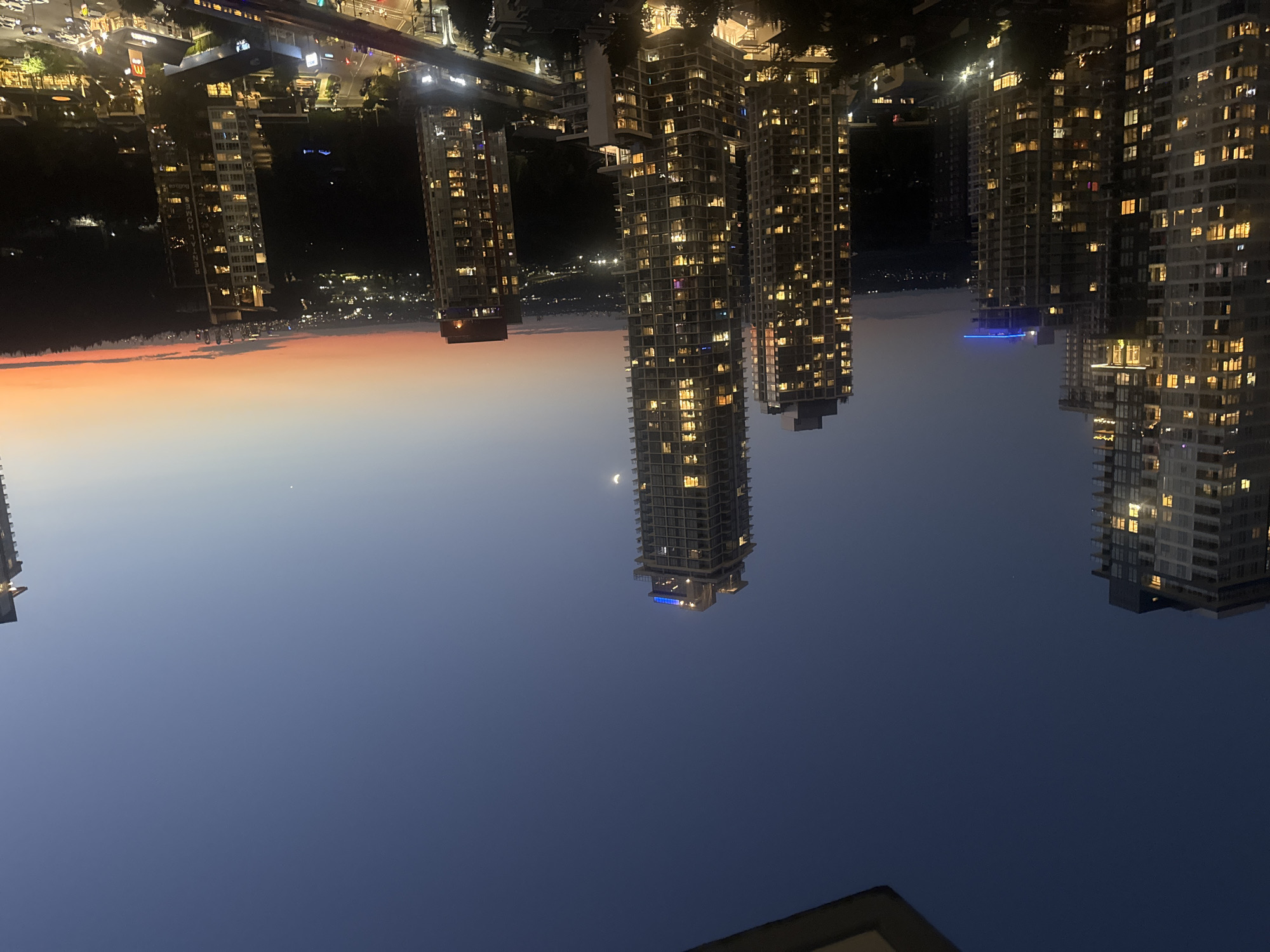 The image depicts a twilight view of the city of Coquitlam, Canada, likely from a high vantage point. Several modern high-rise apartment buildings are prominently featured, illuminated from within by numerous warm yellow lights, indicating that it is evening or night. The sky displays a gradient of colors, transitioning from a deep blue overhead to hues of orange and pink near the horizon, characteristic of sunset or sunrise. A thin crescent moon is visible in the upper part of the sky. Below the buildings, the city lights of Coquitlam stretch out into the distance, creating a sparkling urban landscape. A line of trees forms a dark silhouette against the illuminated city. The foreground appears to be a balcony or rooftop, suggesting the photograph was taken from a residential building. There are no people visible in the image.