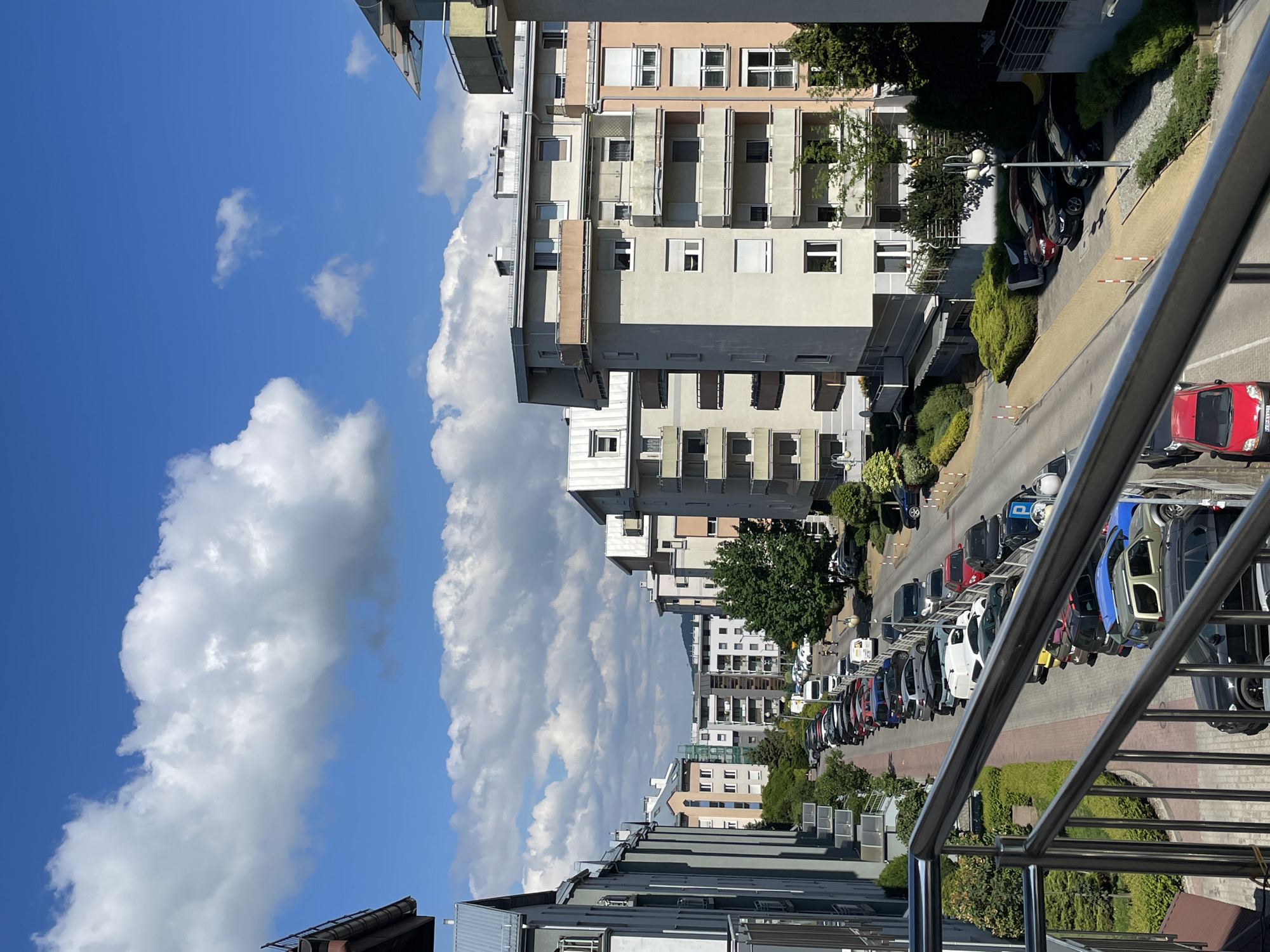 The image depicts a view of a street lined with parked cars and residential buildings under a partly cloudy sky. The scene is observed from an elevated perspective, possibly a balcony, as indicated by the presence of a metal railing in the foreground. The buildings are multi-story structures with a mix of architectural styles and colors, featuring windows and balconies. The street is lined with a variety of cars parked along the side. The sky is blue with large, puffy white clouds. The vegetation includes trees and shrubs along the street. The overall impression is a residential area on a bright day. There is no definitive geographic information to place the location within a specific city.