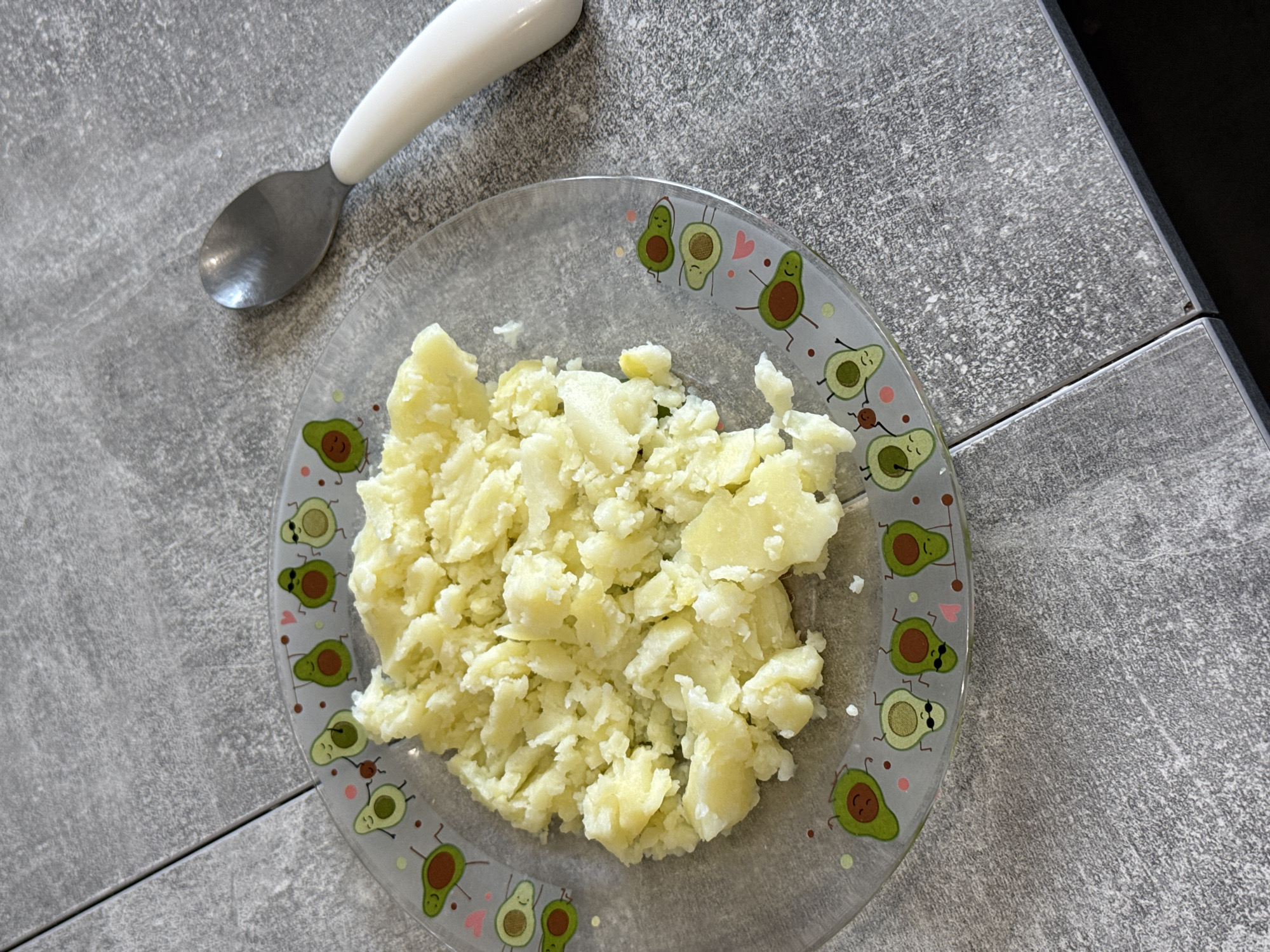 The image shows a close-up, overhead view of a clear glass plate filled with mashed potatoes. The plate has a decorative border featuring cartoon avocados with arms and legs, some wearing sunglasses. To the right of the plate is a silver spoon with a white handle. The plate and spoon are placed on a gray, textured surface that appears to be a tabletop, possibly made of laminate or stone. The lighting suggests it's daytime, possibly indoors. There is no discernible location in the city of Lyubertsy, Russia.