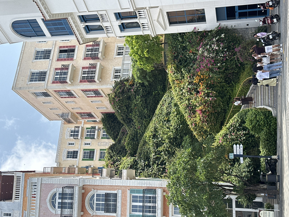 The image captures a detailed view of a street within Sunset Town, Phú Quốc, Vietnam. Three distinct multi-story buildings, exhibiting Mediterranean Revival architecture, frame a central, vegetated incline. The building on the left features a peach-colored stucco facade with arched windows, black iron balconies, and some teal shutters. A light yellow building with green shutters is visible further back and higher up. On the right, a white building presents blue-framed windows and blue shutters with white decorative elements. The central incline is densely covered in green foliage, including various vines and shrubs, intermixed with numerous flowering plants displaying pink, purple, orange, and white blooms. A winding stone staircase with patterned risers and a yellow metal handrail ascends this incline from the foreground, where it is currently occupied by a group of approximately 10-12 individuals. These individuals, mostly young adults dressed in casual summer clothing, are standing on the ground level and the lower steps. Two additional individuals are visible higher up the staircase. On the paved area to the right, three motorbikes are parked. A dark waste bin and a multi-directional signpost with white arrows on a dark pole are located on the left pavement. The scene is illuminated under a partially cloudy sky.