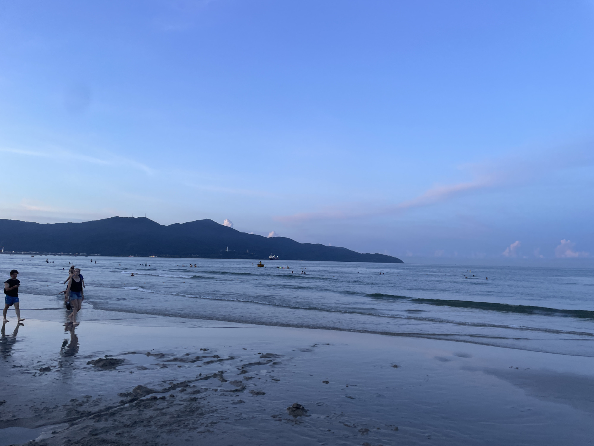 The image depicts a serene beach scene at dusk, with the sky transitioning from a deep blue to lighter shades near the horizon. Silhouettes of mountains rise in the distance, partially obscured by the fading light. The calm ocean water gently laps at the wet sand, creating reflective surfaces that mirror the sky and the figures on the shore.
In the foreground, two individuals, a man and a woman, are walking along the water's edge. The man, wearing a dark t-shirt and blue shorts, is looking towards the left. The woman, in a black tank top and denim shorts, is walking alongside him. They appear to be enjoying a leisurely stroll. Further out in the water, several other people can be seen swimming or wading, their forms indistinct in the dim light. A small yellow boat is visible in the middle distance, adding a touch of color to the scene.
The overall atmosphere is peaceful and tranquil, suggesting the end of a pleasant day at the beach. The soft lighting and the gentle waves contribute to a sense of calm and relaxation. The presence of people enjoying the water and the shoreline indicates a popular public beach. The location cues, such as the mountainous backdrop and the general appearance of the coastline, are consistent with descriptions of beaches in Đà Nẵng, Vietnam. The time of day is clearly evening, as indicated by the twilight sky and the low light conditions. There is no visible text in the image.