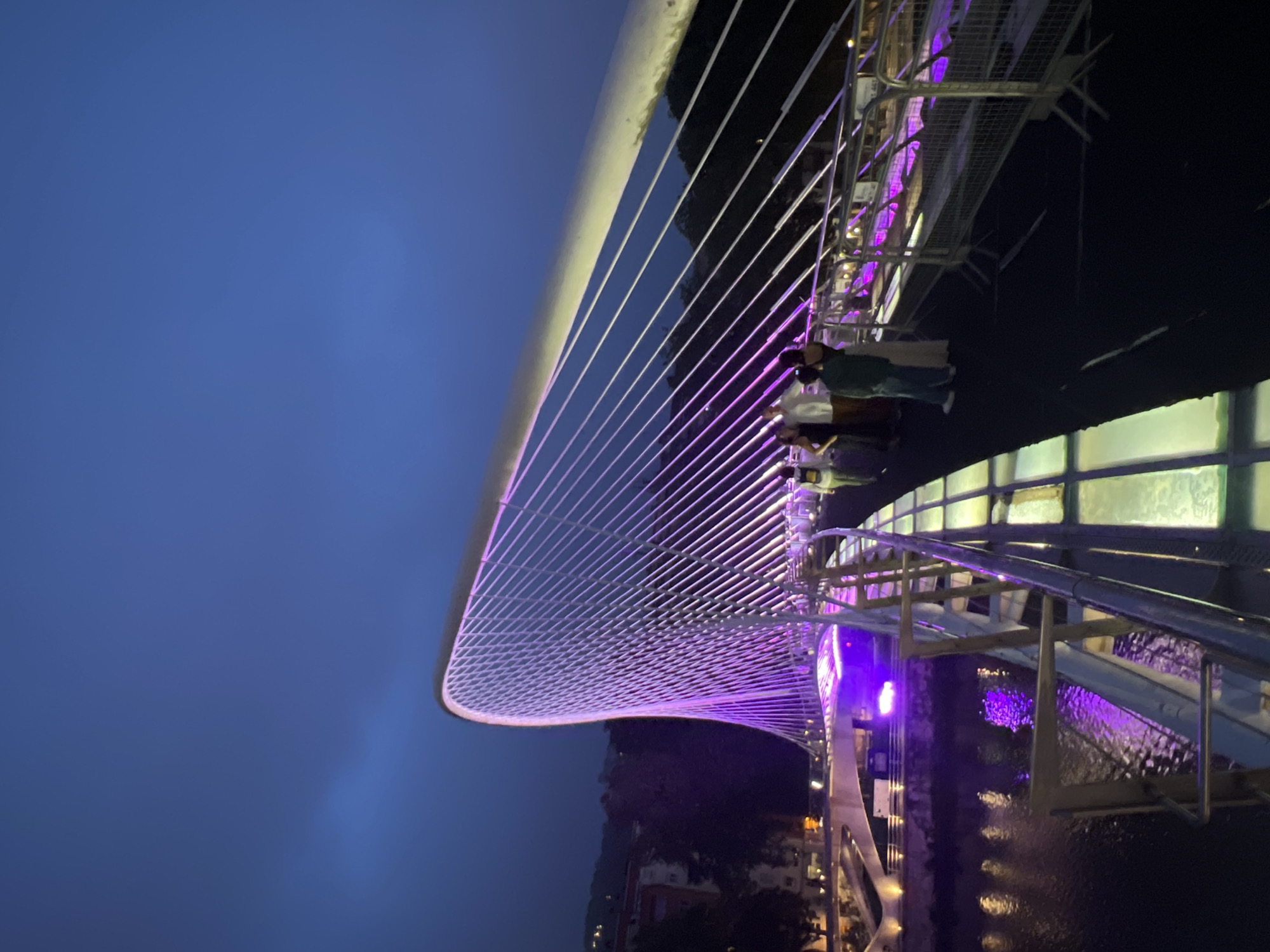 An upward-angle, long shot captures the Zubizuri bridge in Bilbao, Spain at dusk. The iconic white cable-stayed bridge, designed by Santiago Calatrava, is illuminated with vibrant purple and yellow lights, casting a dramatic glow on the scene. The bridge's distinctive sail-like structure rises elegantly against a deep blue twilight sky. Several people, dressed in casual attire, are seen walking across the bridge, their figures silhouetted against the illuminated pathways. The river below reflects the colorful lights, adding to the magical ambiance. The bridge's glass-decked walkway is visible, as are its intricate network of steel cables. The surrounding cityscape is dimly visible in the background, with buildings and trees receding into the darkness. The overall impression is one of modern architectural beauty enhanced by atmospheric lighting and the serene presence of the river.