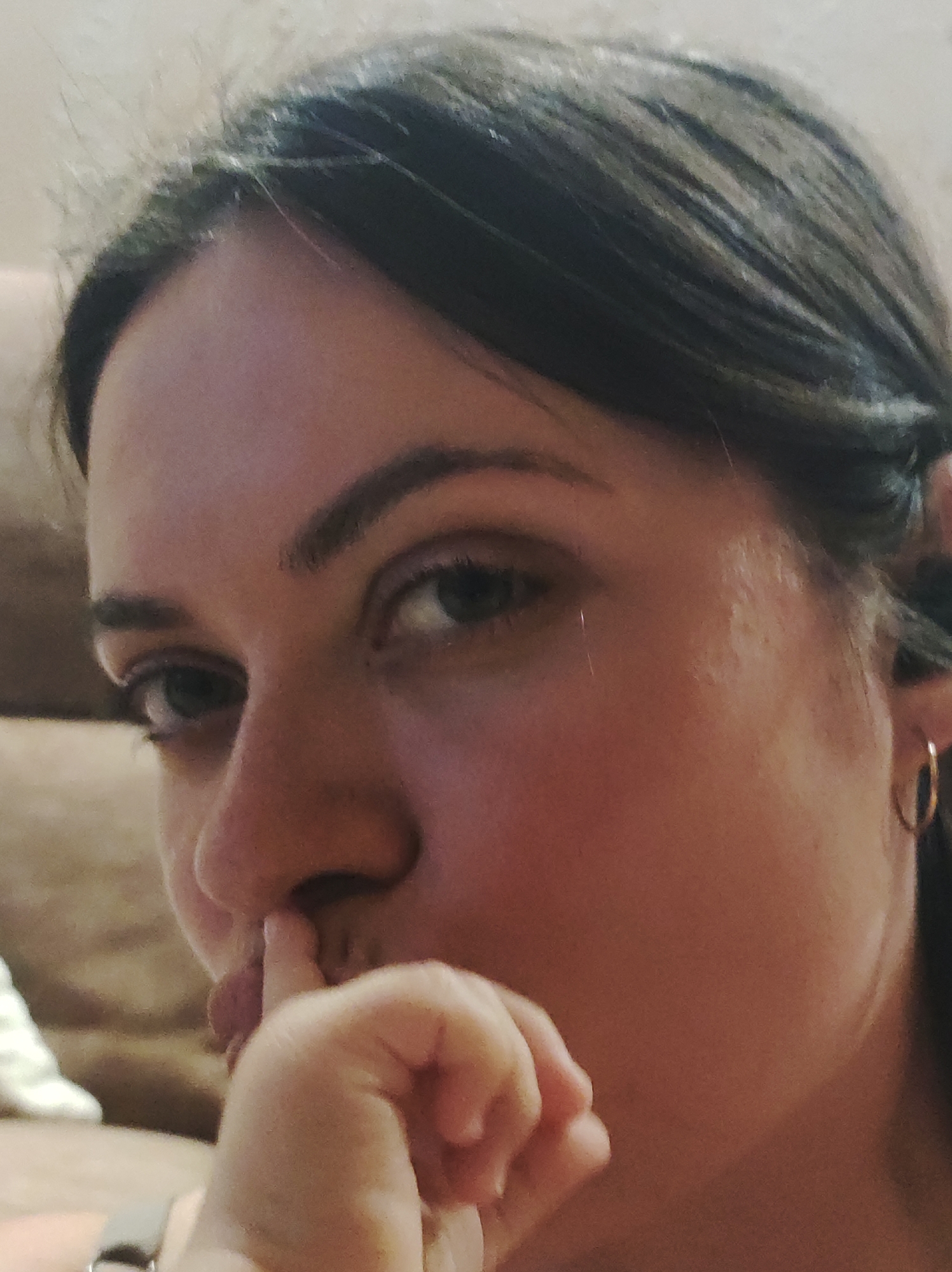 Close-up image of a woman's face. She has dark hair, light skin, and green eyes. Her finger is positioned near her nose and mouth. The background is blurry and indistinct, suggesting an indoor environment. There is no information to suggest the location is in the city of Chaikovsky, Russia.
