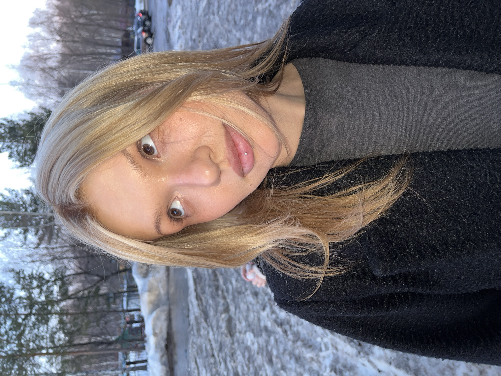 The image shows a young woman with long, blonde hair, light skin, and brown eyes taking a selfie outdoors. She is wearing a dark gray t-shirt and a black textured jacket. She has a piercing on her lower lip. The background shows a snowy landscape with trees and a blurred glimpse of cars in the distance. The scene is likely in a cold, snowy environment. The location is specified as Chaikovsky, Russia.