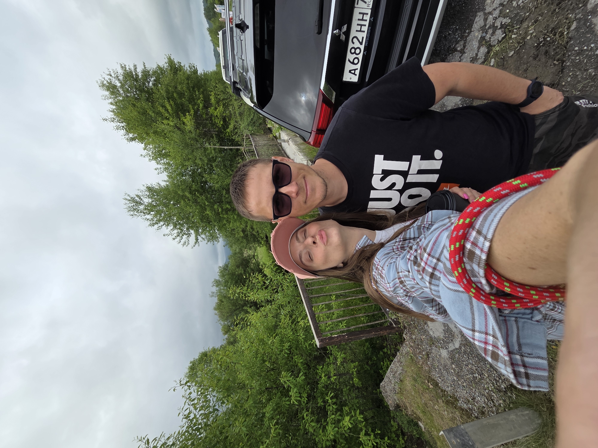 A selfie taken outdoors shows a man and a woman standing next to a black Mitsubishi Outlander. The woman is on the left, wearing a pink baseball cap, a blue and white plaid shirt over a white t-shirt, and her arm is holding the phone. She is puckering her lips towards the camera. The man is on the right, wearing a black t-shirt with "JUST DO IT." printed in white, sunglasses, and camouflage shorts. He has his hand in his pocket. Behind them is lush green foliage, a railing, and a cloudy sky. The license plate of the car reads "A682HH 7". The environment appears to be a roadside with a natural, somewhat undeveloped setting, likely on the outskirts of a town or village. The city of Теплоозёрск, Russia, is a possibility for the location, given the car's license plate region.