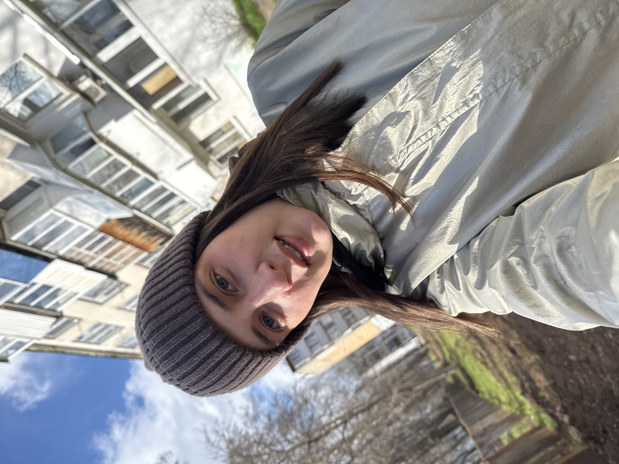 This vertical selfie captures a young woman outdoors in Chaikovsky, Russia. She is positioned in the foreground, looking directly at the camera with a slight smile, revealing her upper teeth. She has long, dark brown hair, blue-grey eyes, and fair skin. She is dressed in a dark grey knitted beanie that covers her forehead and ears, and a shiny, light grey or silver puffer jacket.
The background features a multi-story residential building, likely an apartment complex, characterized by numerous windows and glassed-in balconies, suggesting a functional, perhaps post-Soviet, architectural style. Bare deciduous trees, indicative of late autumn, winter, or early spring, are visible around the building. A patch of green grass can be seen in the lower right. The sky above is bright blue with scattered white clouds, indicating clear, sunny weather. The bright daylight suggests it is midday or early afternoon. The overall atmosphere is casual and relaxed, with the woman appearing content. There is no visible text in the image.