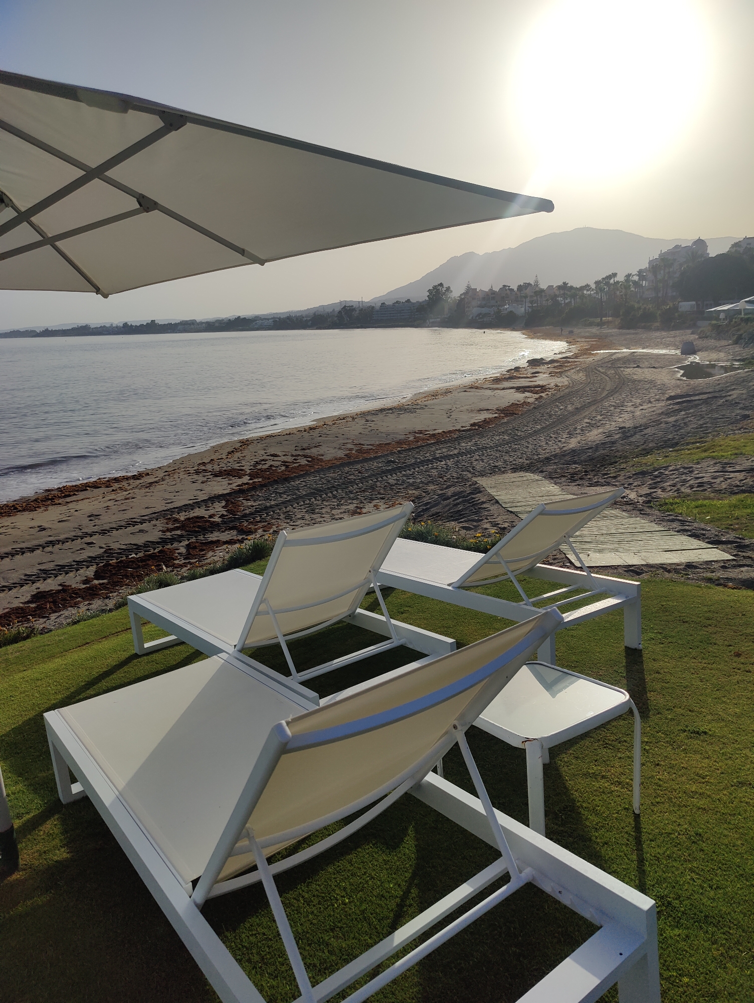 The image shows a beach with the ocean on the left and a sandy shore with scattered seaweed. Two white lounge chairs with beige fabric are set up on a well-manicured green lawn overlooking the beach. A white umbrella is partially visible in the upper left corner, angled over the scene. In the background, mountains rise behind a coastal town with buildings and palm trees. The sun is setting or rising, casting a bright, hazy glow over the landscape. There are no people visible in the image. The overall scene depicts a serene, unoccupied beach resort setting.