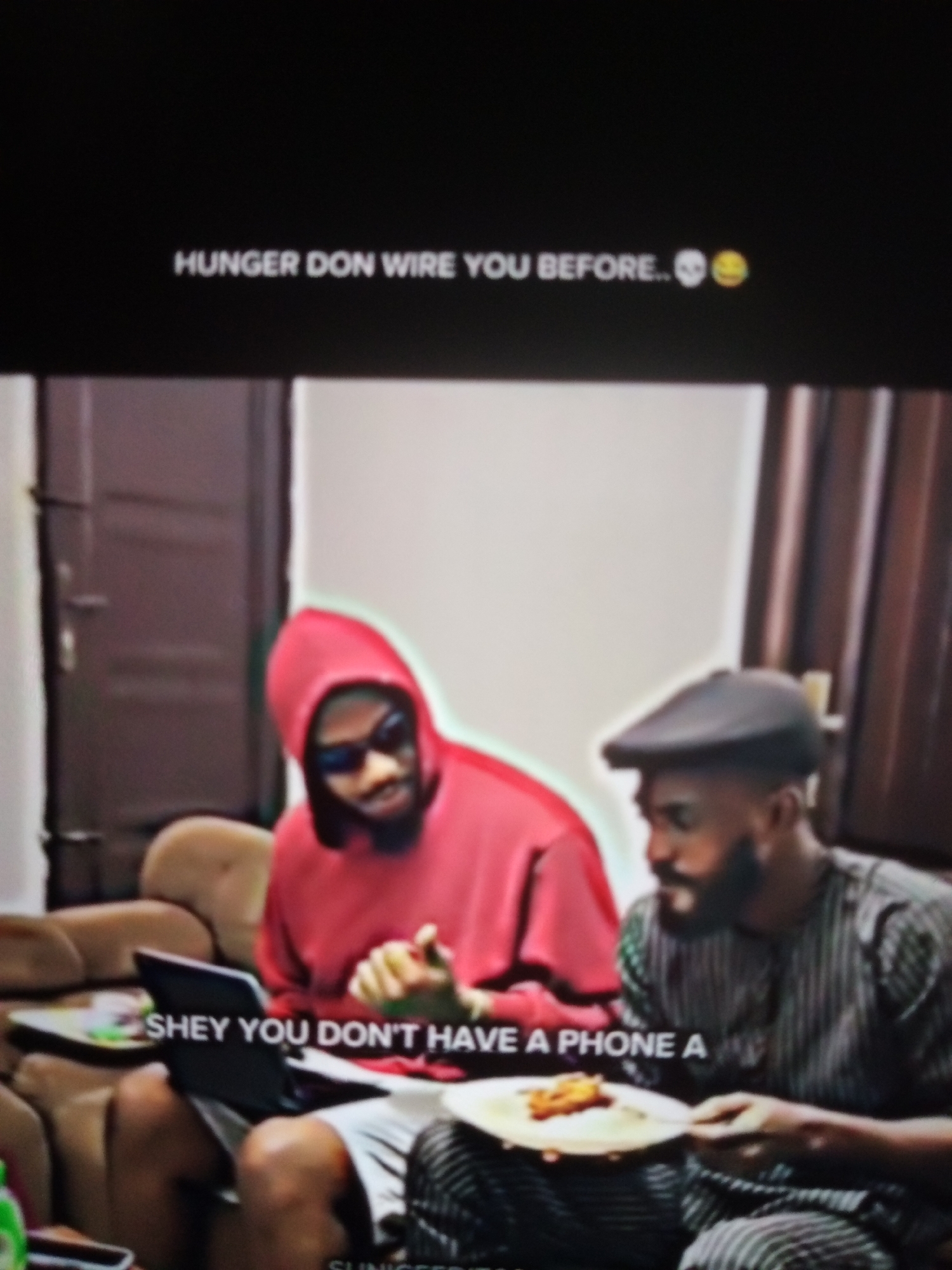 The image shows an indoor scene with two men. The man on the left is wearing a red hooded sweatshirt and sunglasses, and he is seated on a couch. He appears to be holding a laptop or tablet. The man on the right is wearing a dark, striped garment and a cap. He is also seated on a couch and holding a plate of food. Text overlays are present in the image. The upper overlay reads "HUNGER DON WIRE YOU BEFORE.. 💀😂" and the lower overlay reads "SHEY YOU DON'T HAVE A PHONE A". The environment appears to be a living room with couches and a doorway in the background. There is no indication in the image that it is located in Jalingo, Nigeria.