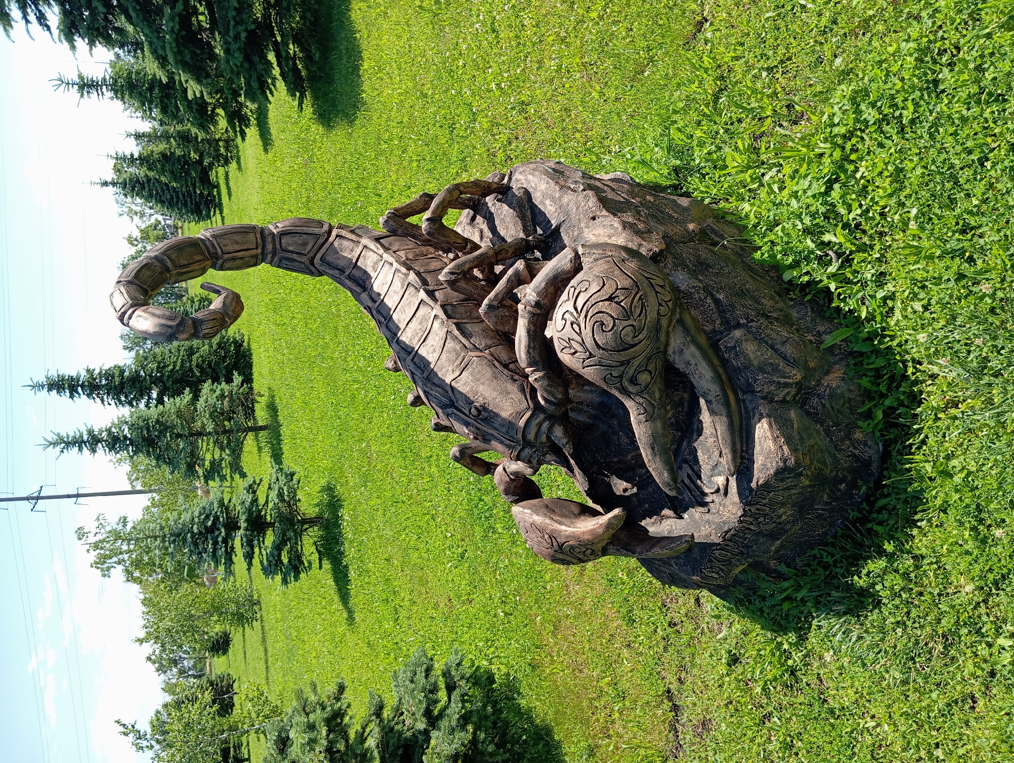 This is a full, eye-level, outdoor, daytime, color photograph of a large, stylized scorpion sculpture in a park in Il'ich, Russia. The scorpion sculpture, made of a brown, textured material that mimics wood or stone, is positioned on a rocky base. It is depicted with its segmented tail curved upwards, and its claws are in front of its body. The sculpture is detailed with intricate carvings, particularly on its claws, which feature ornate scrollwork. The environment surrounding the sculpture is a well-maintained park with vibrant green grass and several coniferous trees of varying sizes. In the background, more trees are visible, along with a clear blue sky and overhead power lines. No people are present in the image. The lighting suggests it is a sunny day.