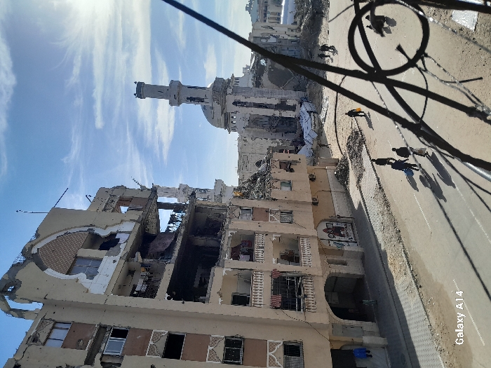 A severely damaged building stands adjacent to a road where several people are walking. The building's structure is compromised, with exposed interiors and missing sections. In the background, a mosque or religious building with a minaret is visible, also appearing damaged. Power lines run across the foreground, partially obscuring the view. The scene depicts a war-torn environment.