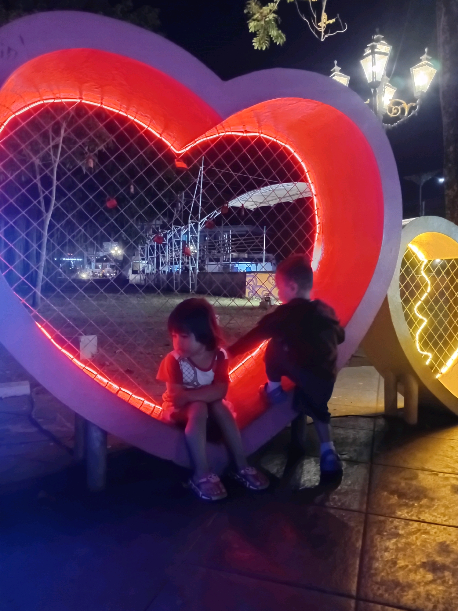In the city of Purworejo, Indonesia, two young children are photographed at night within a large, heart-shaped light installation. The installation is illuminated with bright red and purple neon lights, creating a vibrant spectacle. The children are positioned on a bench inside the heart, which is enclosed by a wire mesh fence. One child, a girl, sits with her arms crossed and her gaze directed downwards. She is wearing a red and white shirt and sandals. Beside her, a boy is leaning forward, appearing to be reaching out or interacting with something beyond the frame. He is dressed in a dark hoodie and shorts, with white socks and blue shoes. The background reveals a dimly lit outdoor environment with trees, streetlights, and what appears to be a fairground or carnival setup in the distance, indicated by scaffolding and red circular decorations. The ground is paved with square tiles, reflecting the colorful illumination from the heart installations.