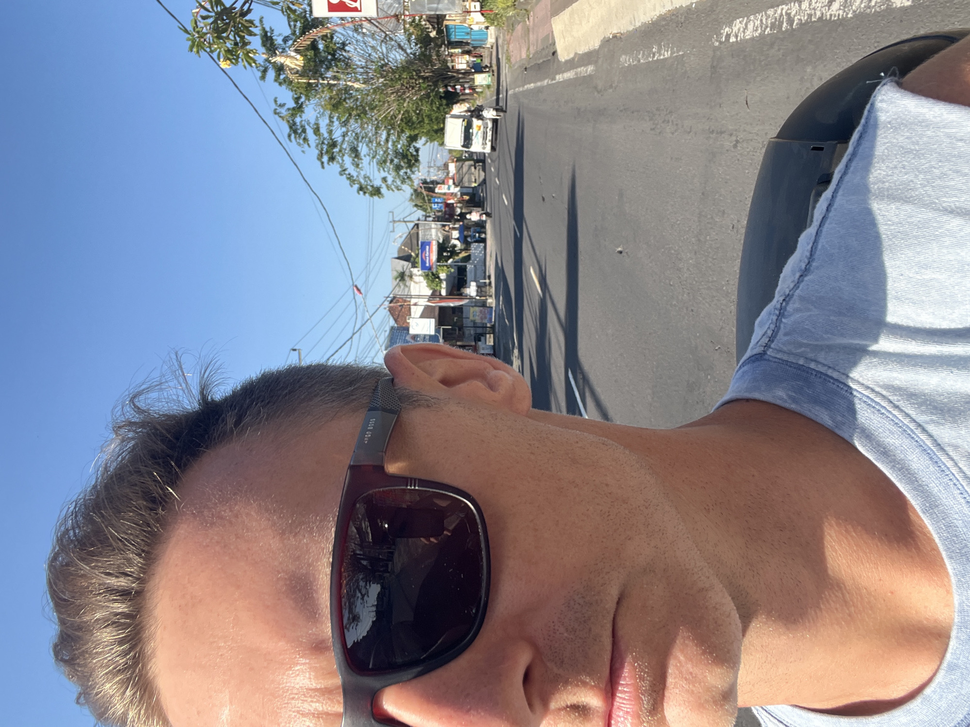 This is a selfie taken from a low angle, showing the face of a person wearing sunglasses and a light blue t-shirt. The person's hair is light brown and slightly messy. The sunglasses are dark-lensed with a red frame and the brand "HUGO BOSS" is visible on the temple. The person is outdoors, with a bright blue sky above and a street scene in the background.
The street is paved and has a white line marking. There are various buildings and shops lining the street, with trees and utility poles. Several vehicles, including a white truck and some motorcycles, are visible on the road, suggesting some traffic. Some of the buildings have signs, one of which appears to be red and white. The scene appears to be a busy commercial area.
The lighting suggests it is daytime, likely mid-morning or afternoon, with the sun casting shadows. The weather appears to be clear and sunny. The context provided indicates the location is Sukawati, Indonesia, which is known for its markets and art. The overall impression is that of a person enjoying a sunny day while on the move through a lively street in Indonesia.