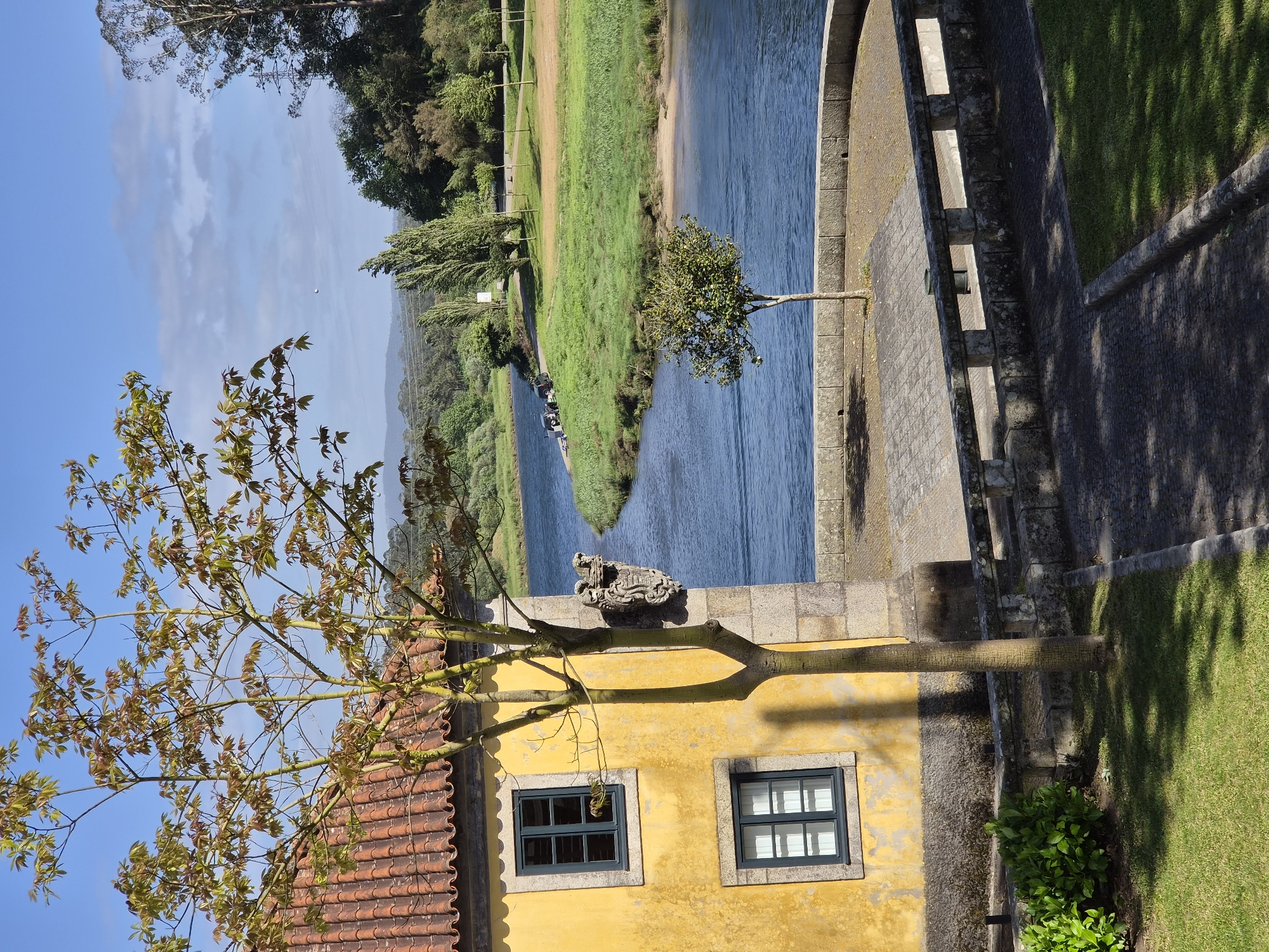 The image captures a scene in Esposende, Portugal. It shows the Cavado River and a portion of the Ponte de D. Luis I bridge. A yellow building with two windows and a red tiled roof is visible in the foreground, with a tree partially obscuring the structure. A stone wall and a grassy area flank the bridge walkway. The river flows through a landscape with trees and grassy banks. The sky is blue with some clouds. There are boats in the river.