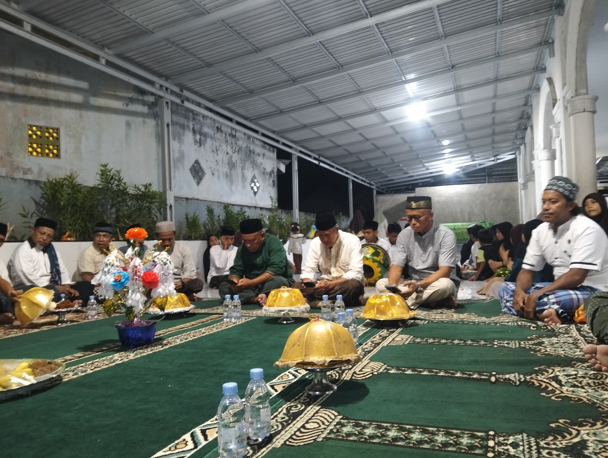 The image depicts an outdoor gathering at dusk, possibly for a religious or community event, set in Petapa, Indonesia. The scene is characterized by a large, covered open-air space with a corrugated metal roof supported by metal beams. The floor is covered with a green carpet featuring an intricate cream and black patterned border.
A group of people, primarily men, are seated on the carpet, facing towards the viewer's left. They are dressed in traditional Islamic attire, including peci (caps) and long-sleeved shirts or tunics. Many are wearing peci caps of various designs and colors, including black, white, and patterned ones. Some individuals are wearing sarongs.
In the foreground, several individuals are visible. To the left, a man in a white shirt and black peci is seated. In the center, a decorative floral arrangement with orange, blue, and red flowers in a blue vase is placed on the carpet. Several golden, dome-shaped objects are also prominently displayed on pedestals on the carpet, some appearing to be decorative or ceremonial. Bottles of water are arranged on the carpet near the seated individuals, suggesting refreshments are being served.
In the background, more people are visible, including some women and children. The walls in the background appear to be concrete, with some decorative elements like a square patterned vent on the left wall. The lighting suggests artificial illumination from overhead fixtures, creating a somewhat dim ambiance typical of an evening event. The overall impression is one of a communal gathering or ceremony taking place in a semi-public, covered outdoor space.