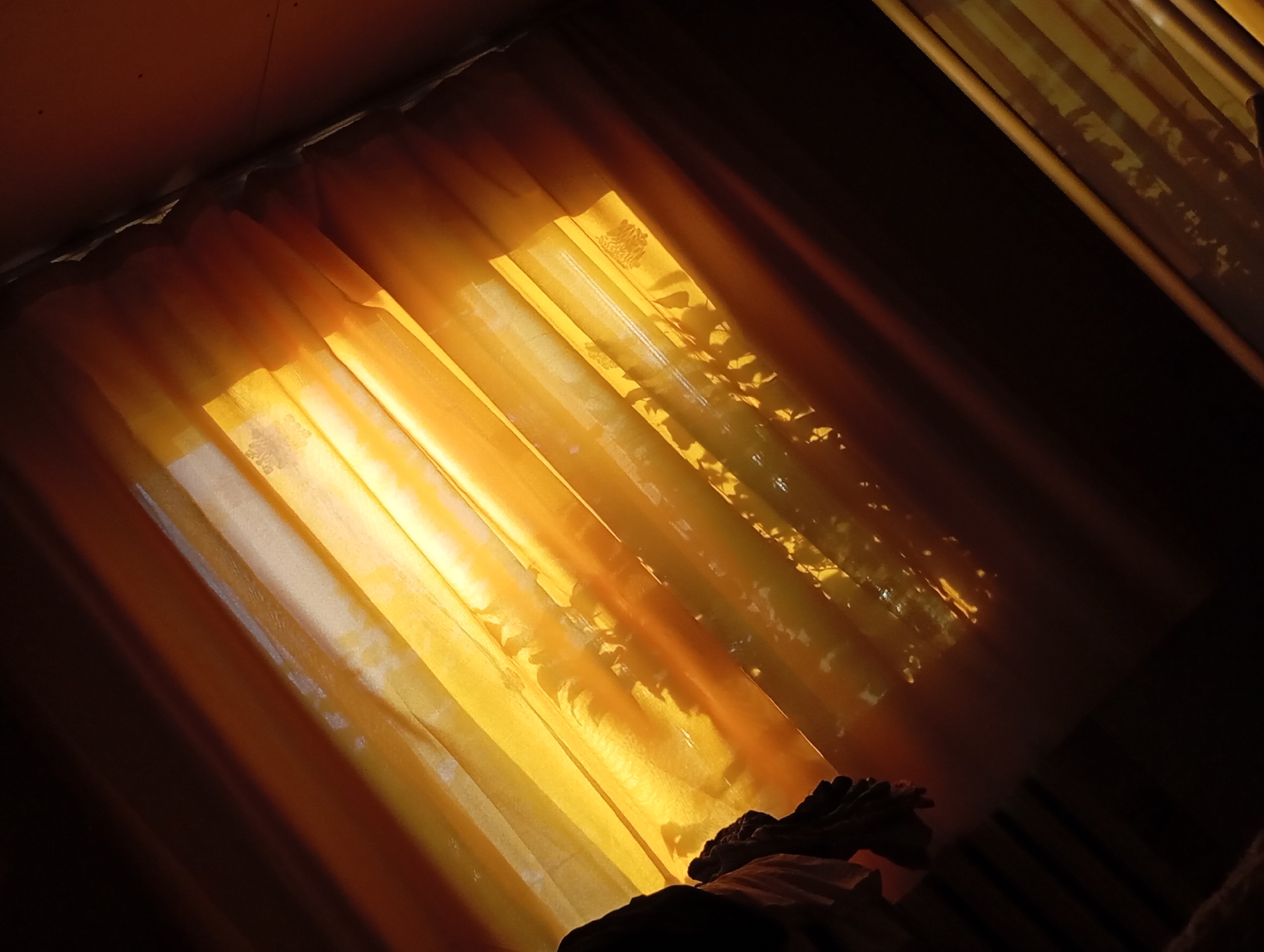 The image shows a window with golden yellow curtains, partially open, allowing light to stream into a dark room. The light creates a bright, slightly blurred effect, with shadows of foliage visible on the curtains. In the lower part of the frame, there is a dark shape that appears to be a person's head and shoulders resting on a bed, partially covered by a sheet. The scene is dimly lit, with the primary light source being the window.
I cannot determine the city of Фоки, Russia from the provided image.