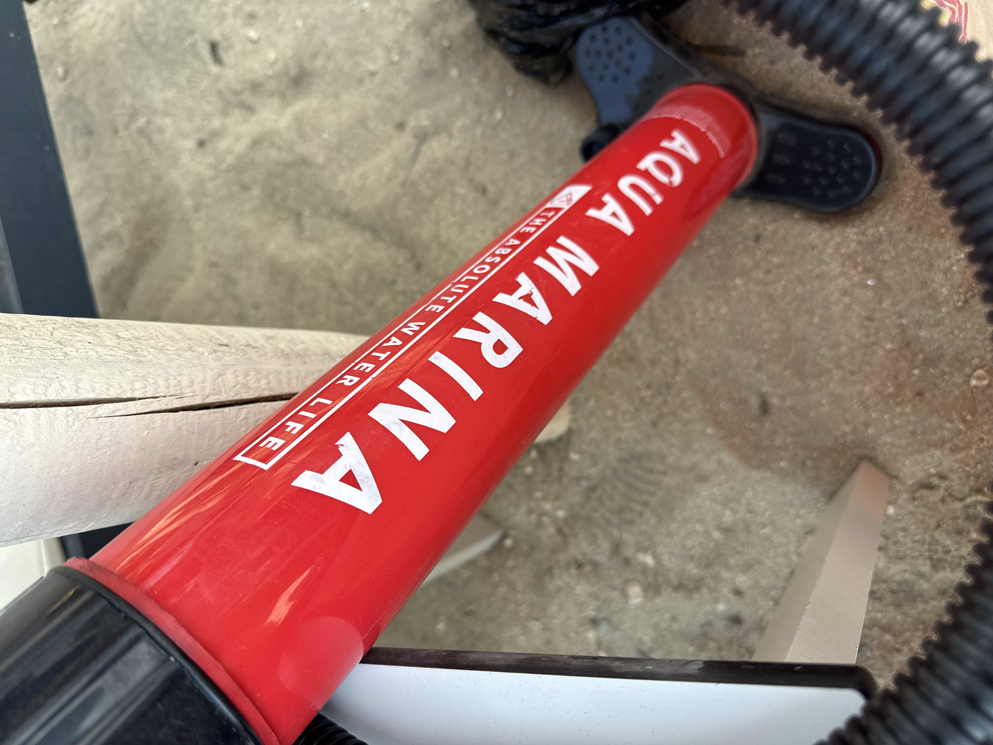 A close-up, oblique shot shows a red pump with white text on it. The text reads "AQUA MARINA THE ABSOLUTE WATER LIFE." The pump is positioned diagonally, with the handle end pointing towards the top left of the frame. A black ribbed hose is attached to the bottom of the pump. The background consists of sand and a light-colored wooden post. The scene appears to be outdoors, possibly near a beach. The sand has subtle footprints visible. The lighting is bright, suggesting daylight. There are no people or animals visible in the image.