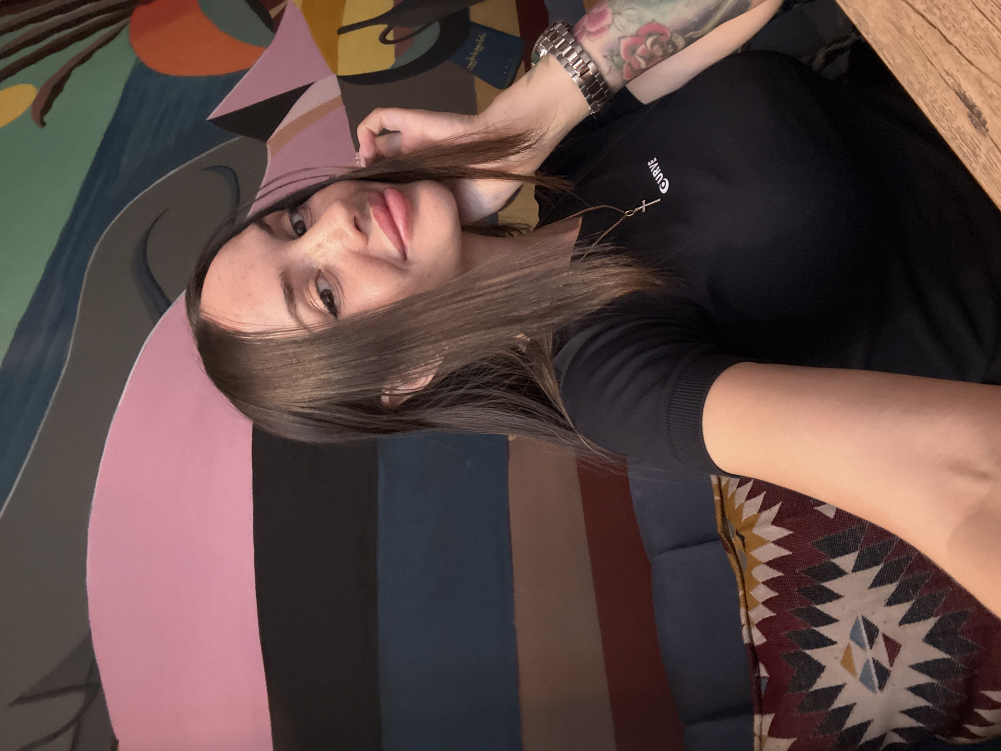 A young woman with brown hair and freckles is taking a selfie in what appears to be a cafe or restaurant. She is wearing a black t-shirt with a cross pendant and a watch on her left wrist. The background is a colorful mural with abstract shapes and stripes. A patterned pillow is visible on a blue couch to her left. The overall impression is a casual and stylish setting. The presence of a watch and the woman's attire do not suggest any specific location within Saint Petersburg, Russia, or any other city.