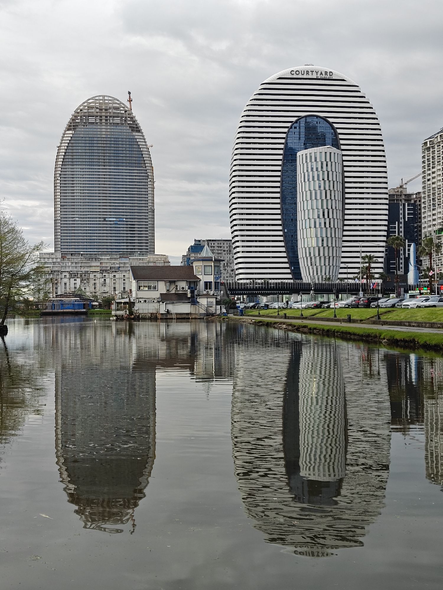 The image presents a modern cityscape viewed across a tranquil body of water under an overcast sky. Two prominent high-rise buildings dominate the scene, flanked by smaller structures and a lively waterfront.
On the left, a tall, slender, and oval-shaped skyscraper is under construction, with a visible skeletal framework at its rounded top and a construction crane. Its facade is primarily glass, reflecting the muted light of the cloudy sky.
To its right stands the distinctive horseshoe-shaped "COURTYARD BY MARRIOTT" hotel. This building is characterized by bold horizontal black and white stripes and a large central arch. Within this arch, a smaller, cylindrical building with a patterned white and grey facade is nestled, surrounded by dark blue glass.
Between these two skyscrapers and closer to the water, several smaller, older-style buildings with pitched roofs and a more traditional appearance are visible, including one with a small turret. The immediate shoreline is green and well-maintained, with a path running alongside a row of parked cars. Flags, including what appears to be the flag of Georgia (red cross on a white background), are visible near the Courtyard by Marriott building.
The calm surface of the water perfectly reflects the buildings, creating a symmetrical yet slightly distorted mirroring of the urban landscape due to subtle ripples. The sky is uniformly cloudy, casting a soft, diffused light over the entire scene.