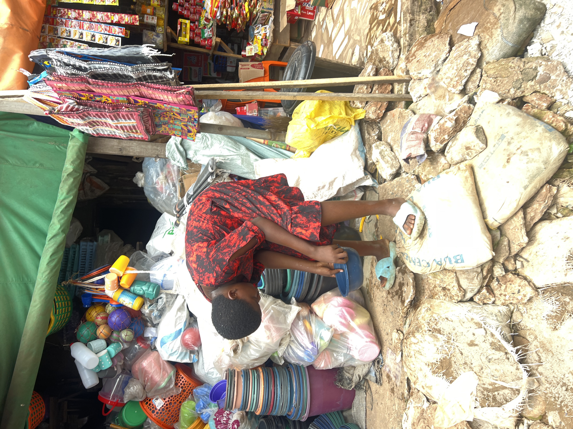 This is a medium shot of a young person, likely a child, in a market in Nkpor, Nigeria. The person is wearing a red and black patterned dress and is partially lying down, surrounded by various goods. The scene appears to be a market stall with a green tarp covering one side. In the foreground, there are stacks of colorful plastic bowls and containers, as well as some plastic balls and bottles. Behind the person, there are bags of what appears to be cement, as well as piles of rocks and debris. In the background, there are shelves stocked with various products, including packaged goods and what look like small sachets of food items. The lighting suggests it is daytime, and the environment appears to be outdoors, possibly on a dusty or unpaved surface given the rocks and dirt. The overall impression is one of a busy, informal market setting. The person's posture and engagement with the objects suggest they might be interacting with the merchandise or taking a rest amidst the commerce. There is text visible on one of the white bags that appears to say "BURCEM". The mood is functional and captures a slice of everyday life in a Nigerian market.